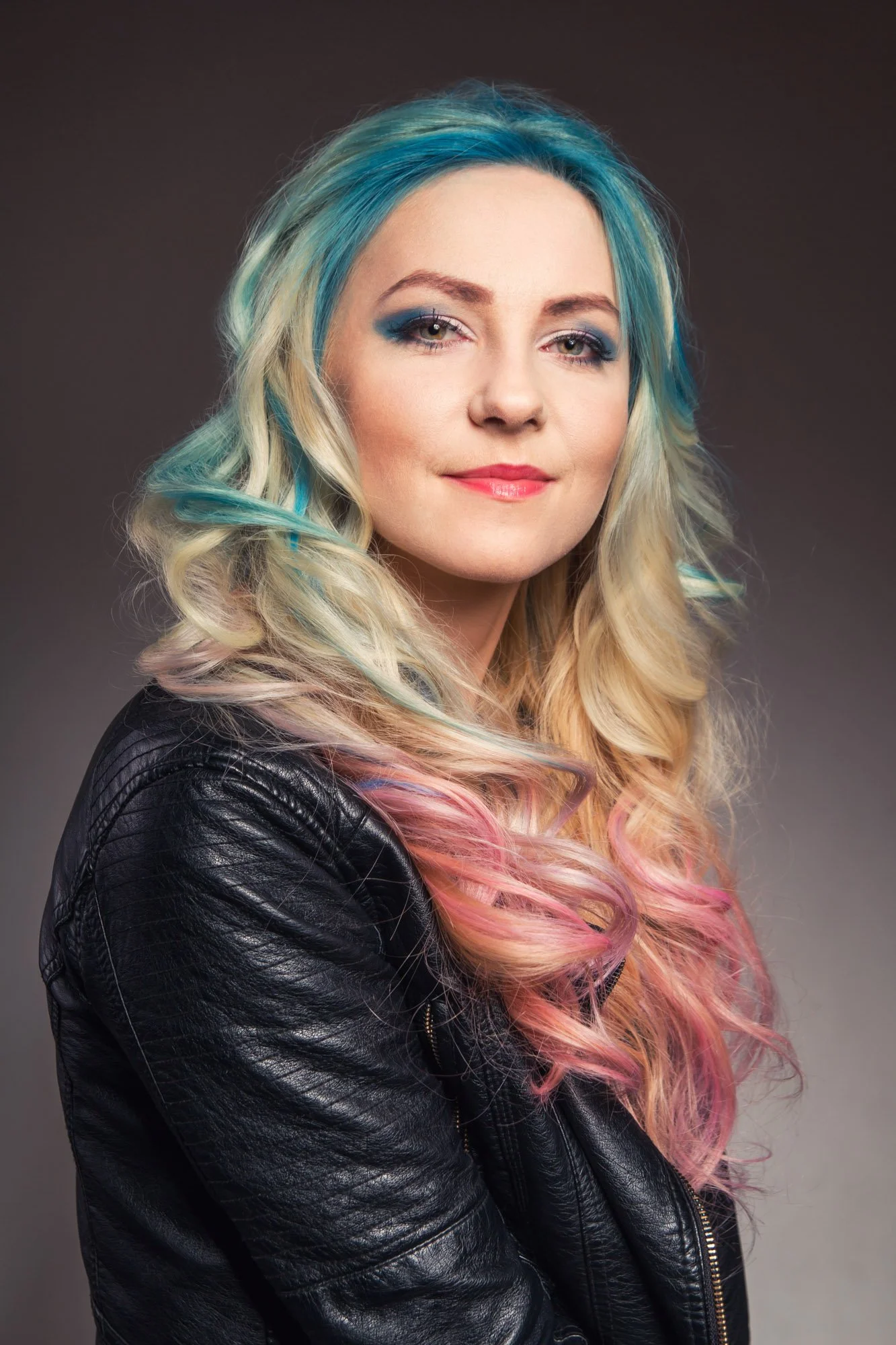 Portrait, Fitness & Commercial Photographer Zurich. A woman with blonde hair featuring blue and pink highlights, wearing a black leather jacket, is smiling slightly against a dark gray background.