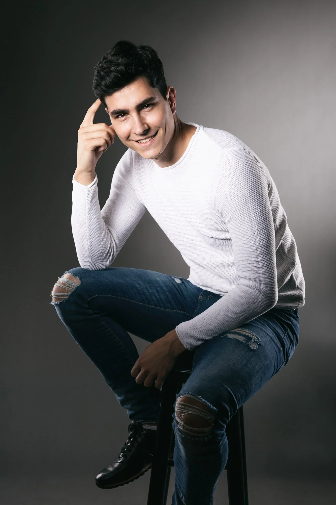 Portrait, Fitness & Commercial Photographer Zurich. Portrait of a smiling young man with dark hair, wearing a white long-sleeved shirt and torn jeans, sitting on a stool against a gradient background.
