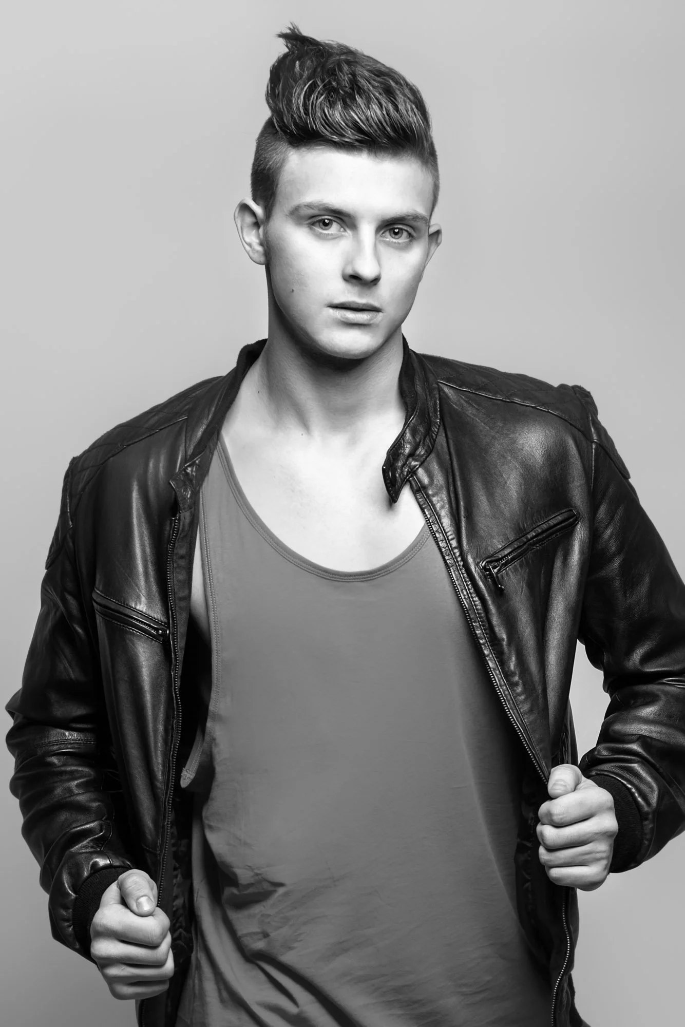 Black-and-white portrait of a young man with styled hair, wearing a leather jacket over a T-shirt, looking directly at the camera with a serious expression, against a plain background.