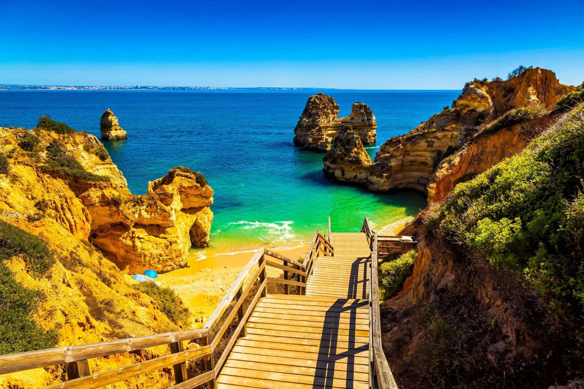 A wooden staircase leads down to a sandy beach with a parasol and towel. The beach is surrounded by tall, rocky cliffs covered with vegetation, overlooking the ocean with clear blue water. In the background, there are more rocky formations and a dist