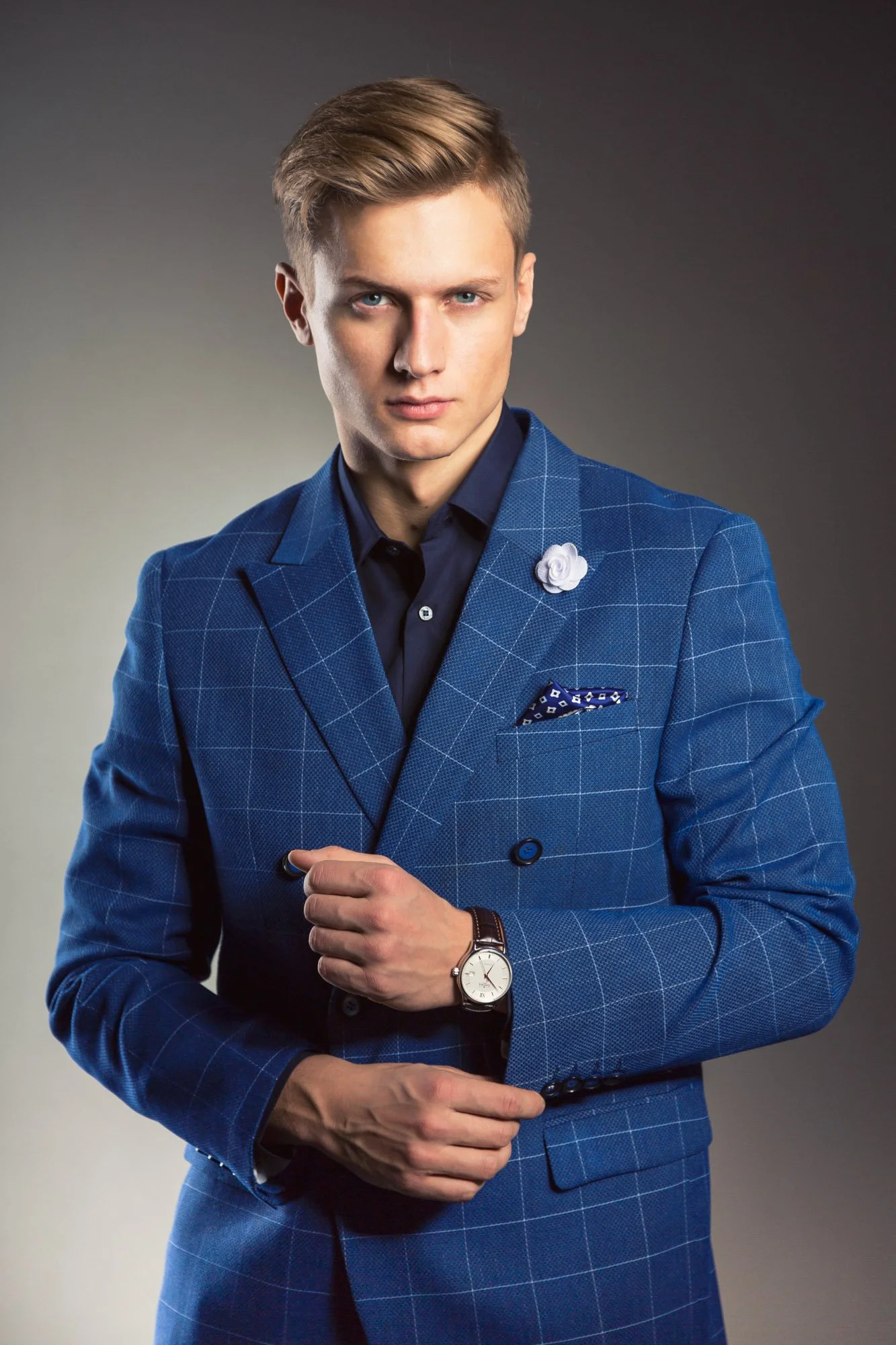 Portrait, Fitness & Commercial Photographer Zurich. A young man in a blue checkered suit, dark shirt, and a boutonnière, standing against a gradient gray background, with his right hand adjusting his coat sleeve and wearing a watch.