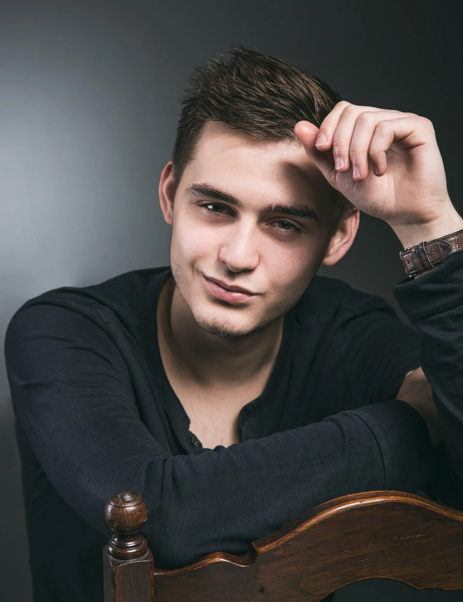 A young man with styled brown hair, light skin, and blue eyes sitting behind a wooden chair with round finials, wearing a black long-sleeve shirt and a wristwatch, leaning slightly forward with one hand touching his forehead, against a dark gray back