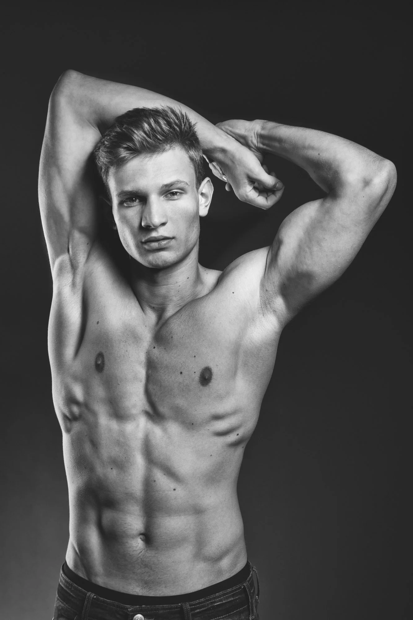 Portrait, Fitness & Commercial Photographer Zurich. Black and white photo of a shirtless young man with short hair, flexing behind his head with one arm, looking at the camera against a plain dark background.