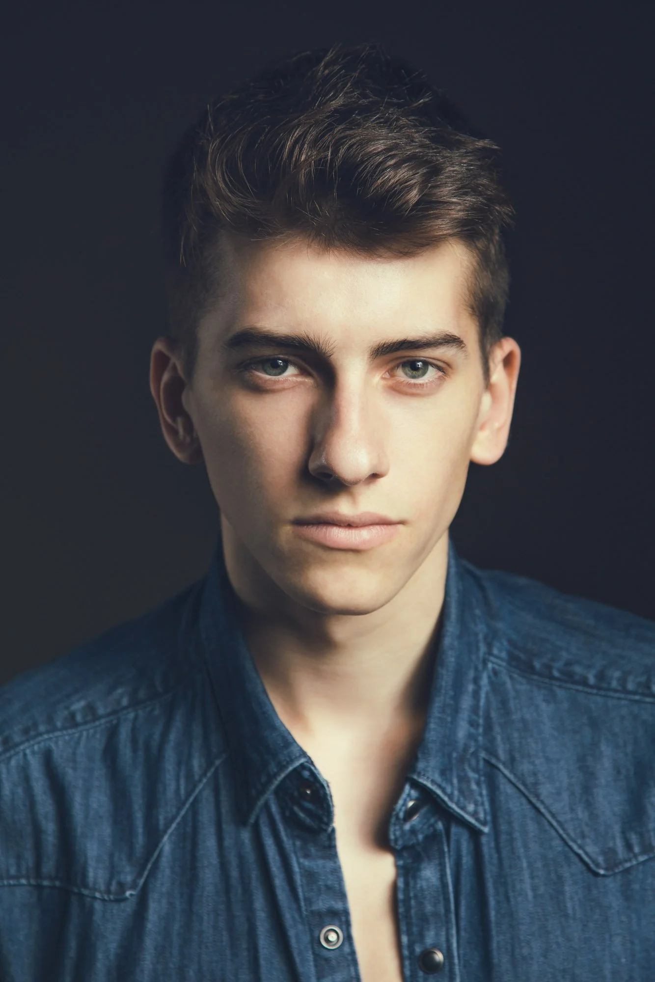 Portrait, Fitness & Commercial Photographer Zurich. Close-up of a young man's face with fair skin, short brown hair, looking directly at the camera against a dark background, wearing a denim shirt.