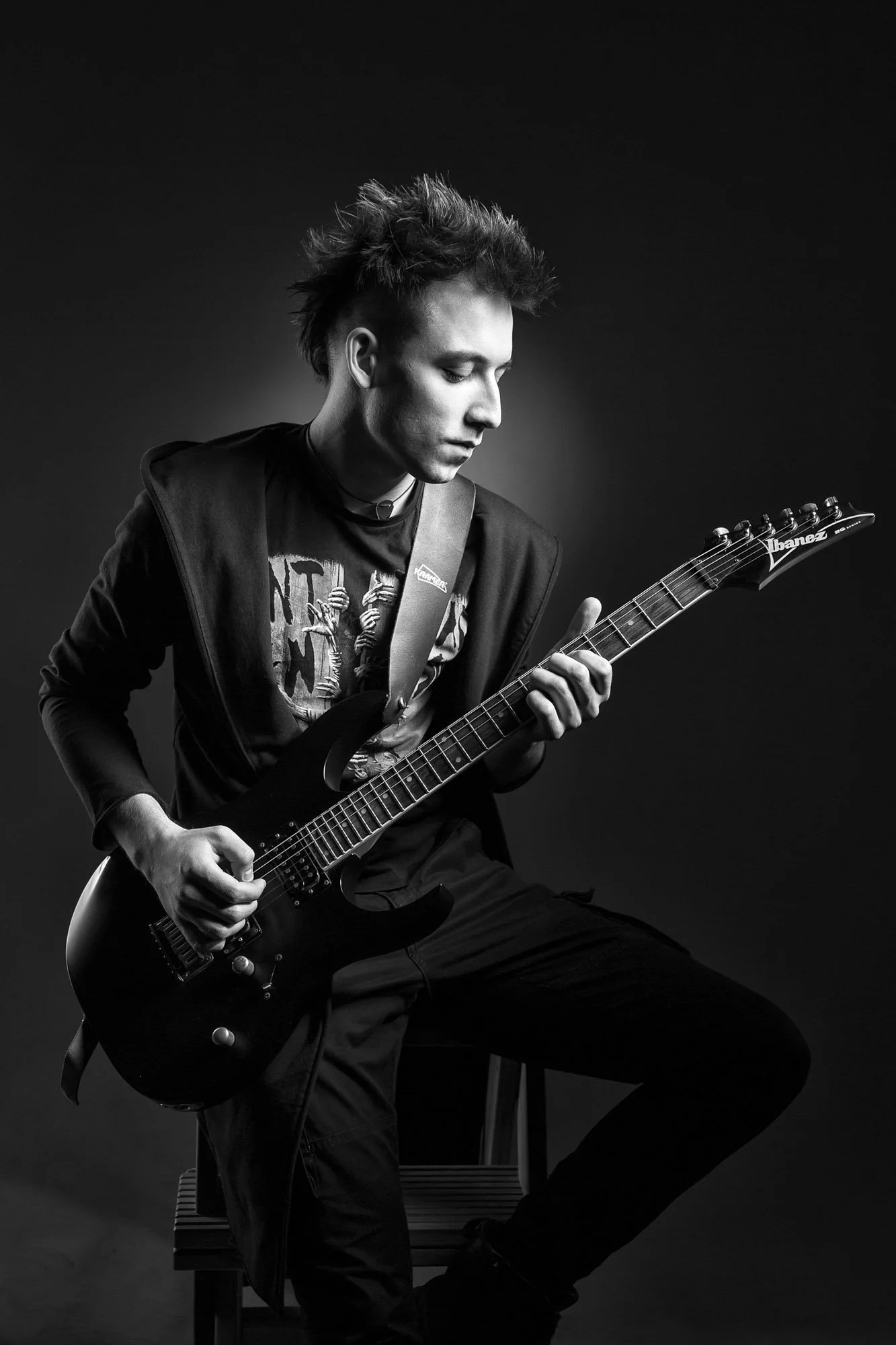 Black and white photo of a young man with spiked hair playing an electric guitar, sitting on a stool against a dark background.