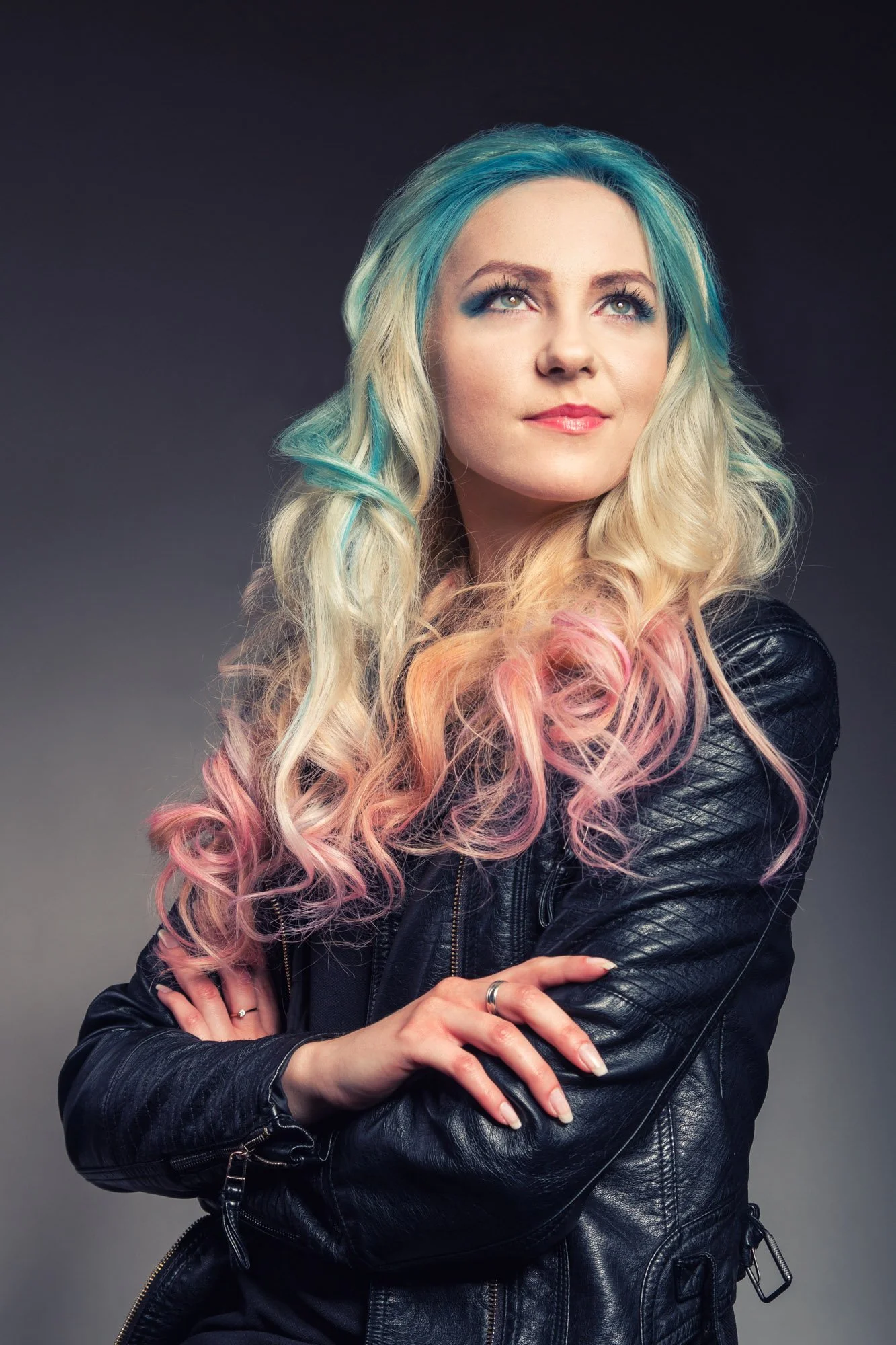 A woman with long, wavy hair dyed in blue, blonde, pink, and peach colors. She is wearing a black leather jacket, has crossed arms, and is looking slightly upward with a confident expression against a dark gradient background.