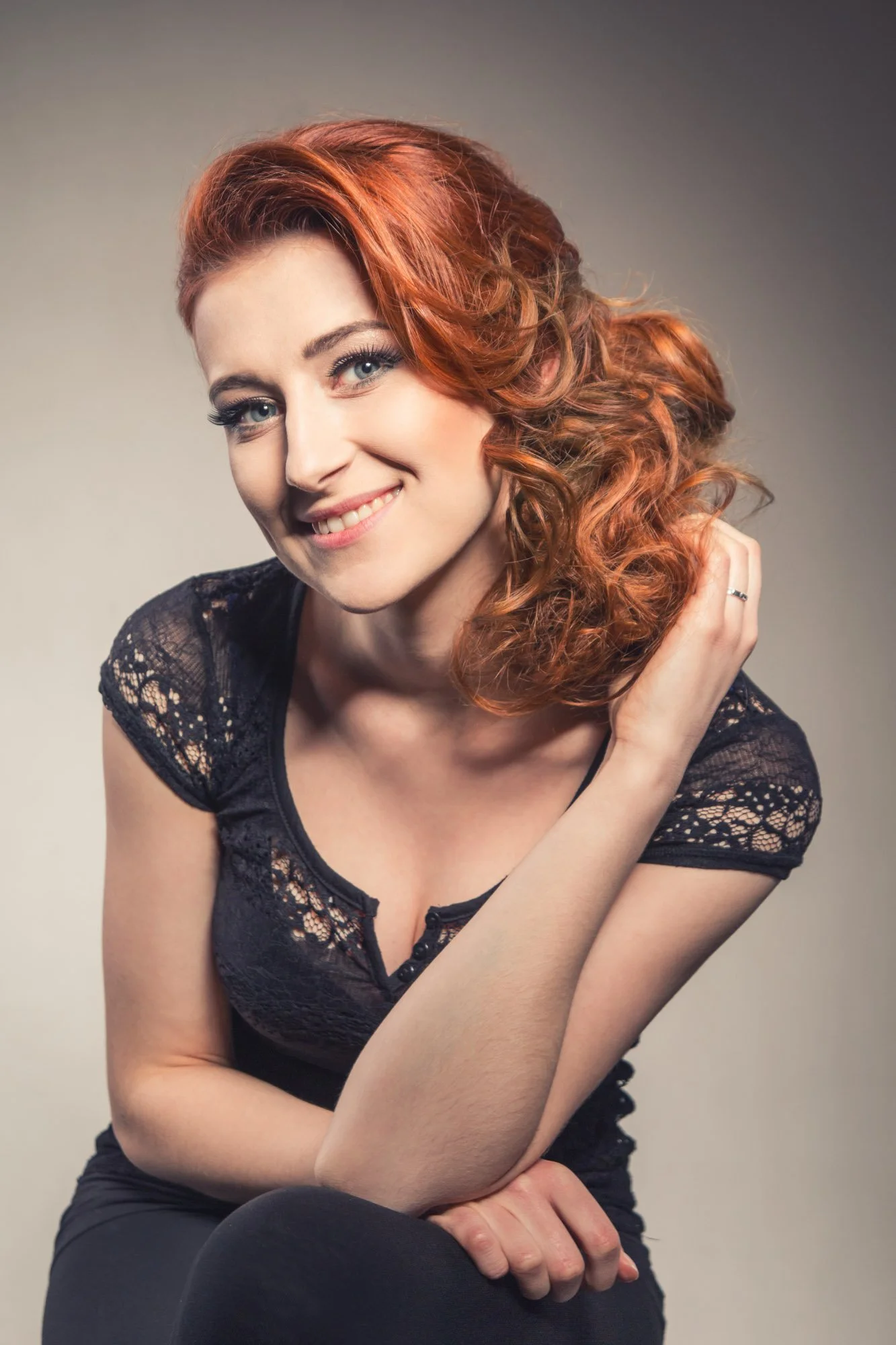 A woman with red, curly hair wearing a black lace top, smiling, sitting with arms crossed, against a neutral background.