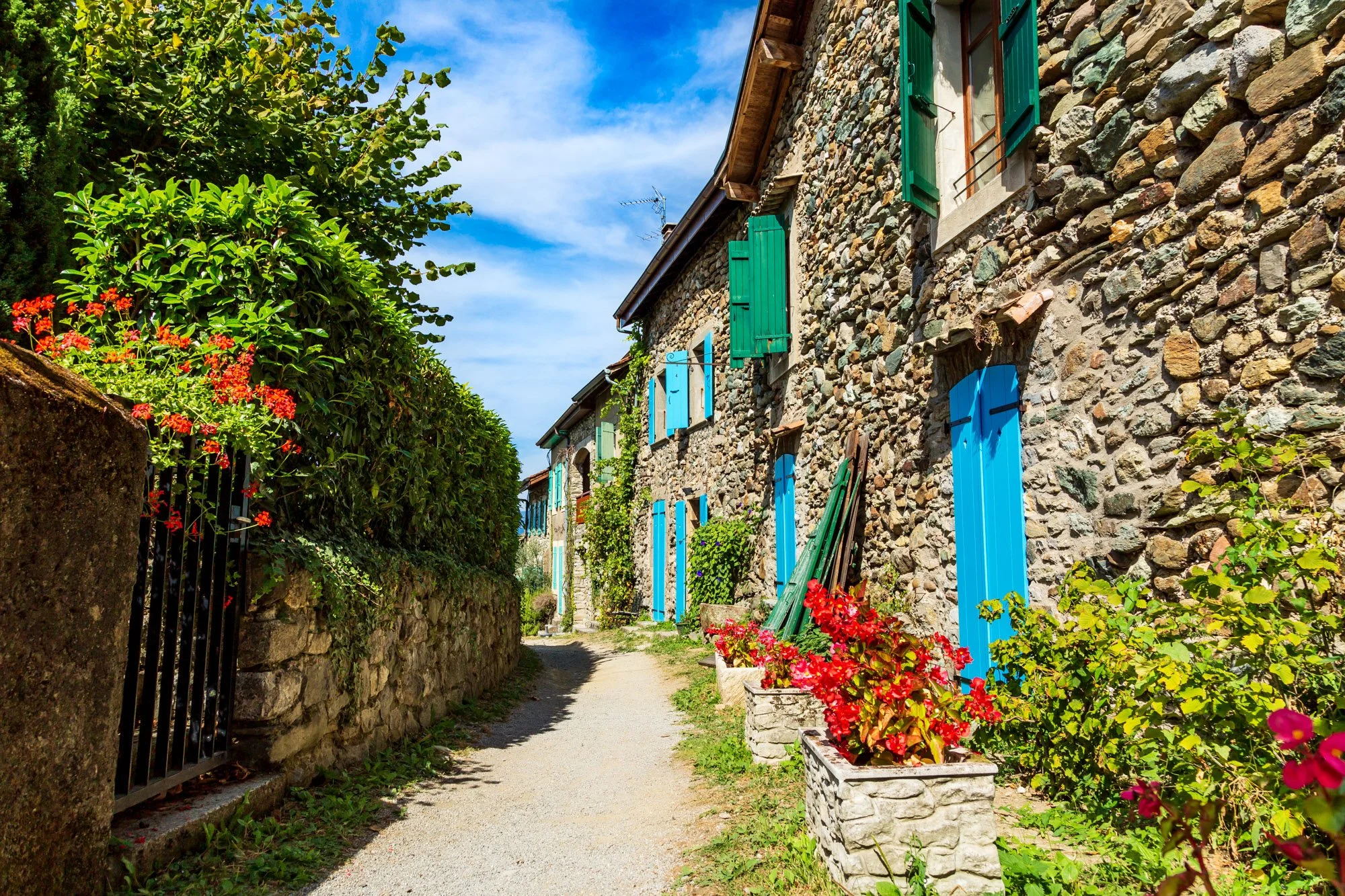 Fine art print photogA narrow dirt pathway runs alongside a stone house with colorful open shutters on the windows. Flower pots with vibrant red and pink flowers line the path. A lush green hedge is on the left, and the sky is clear with some clouds.
