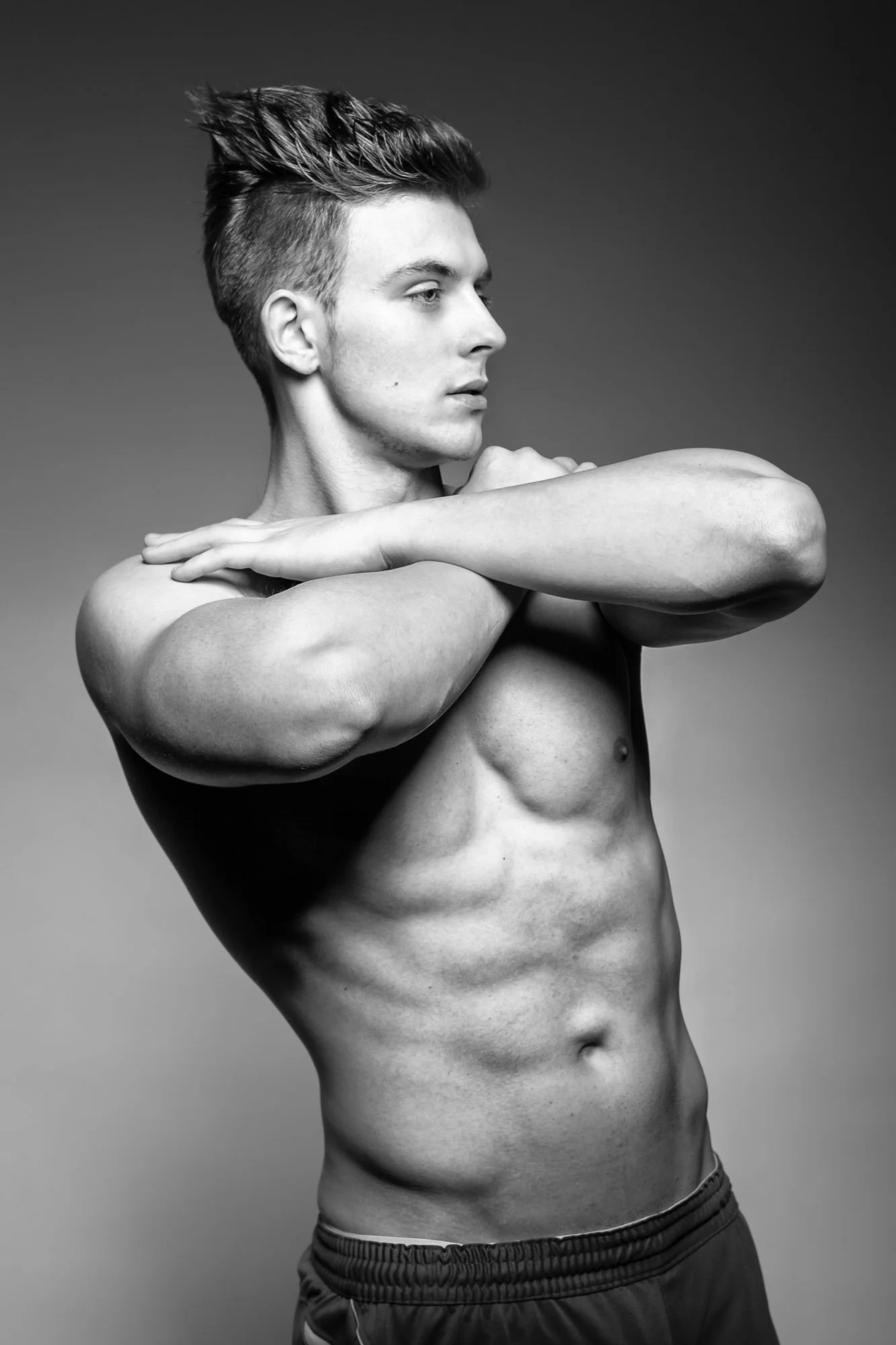 Black and white photo of a muscular man with short styled hair, shirtless, crossing his arms with one hand on his shoulder, in a studio setting.