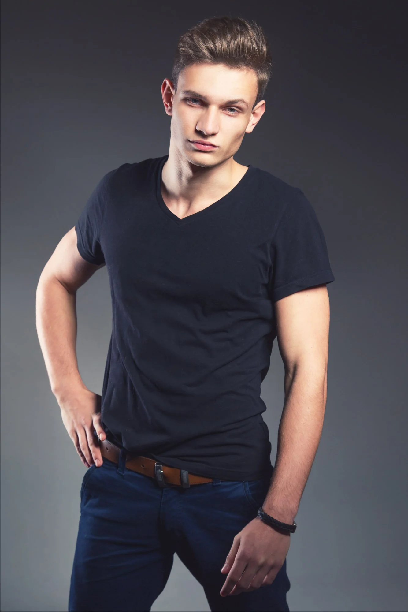 Artistic male portrait photography Zurich. A young man with short brown hair, wearing a black V-neck T-shirt and dark pants, posing with one hand on his hip against a gray background.