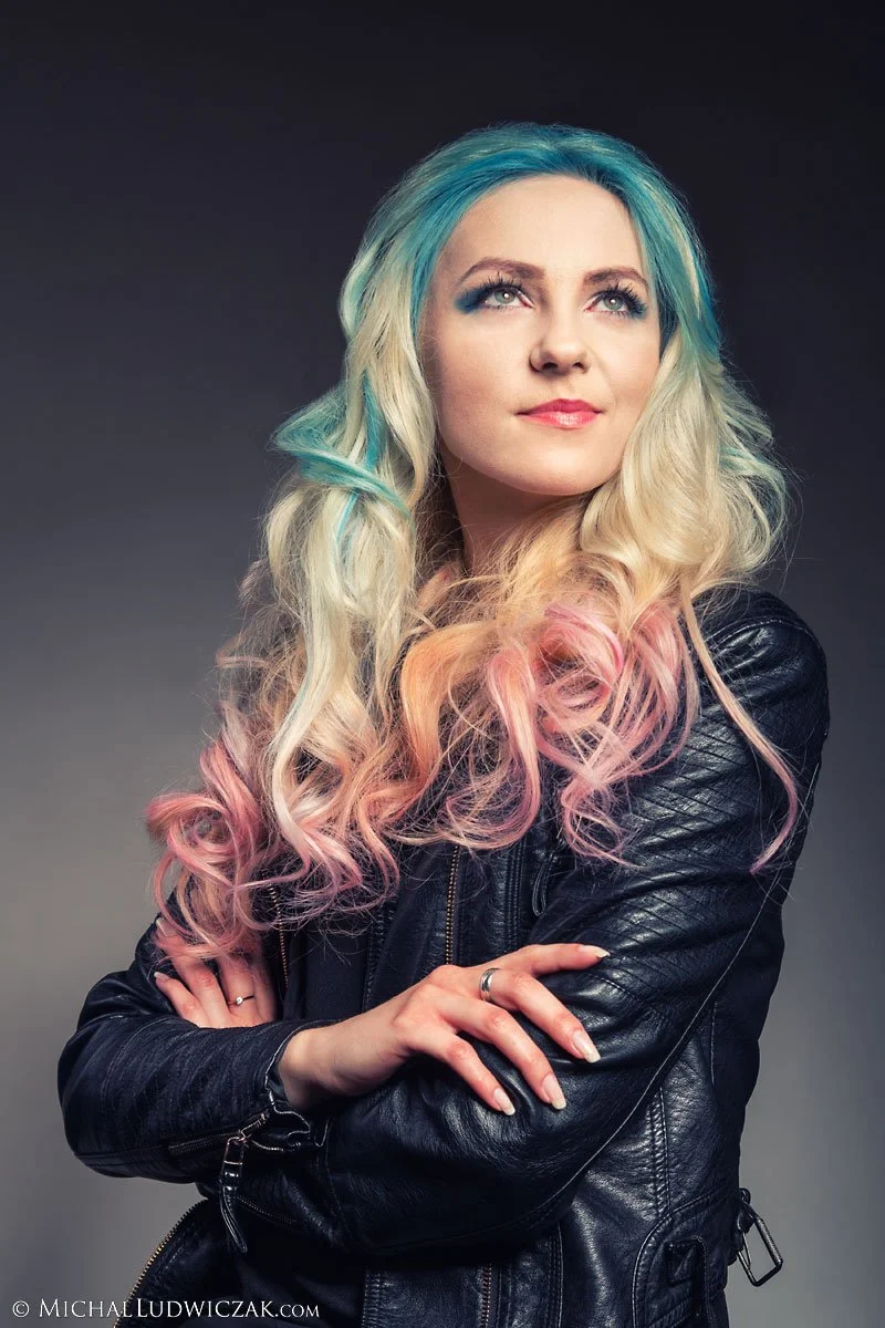 Portrait of a woman with long, wavy hair dyed in pastel shades of blue, pink, and blonde, wearing a black leather jacket, posing against a dark background.
