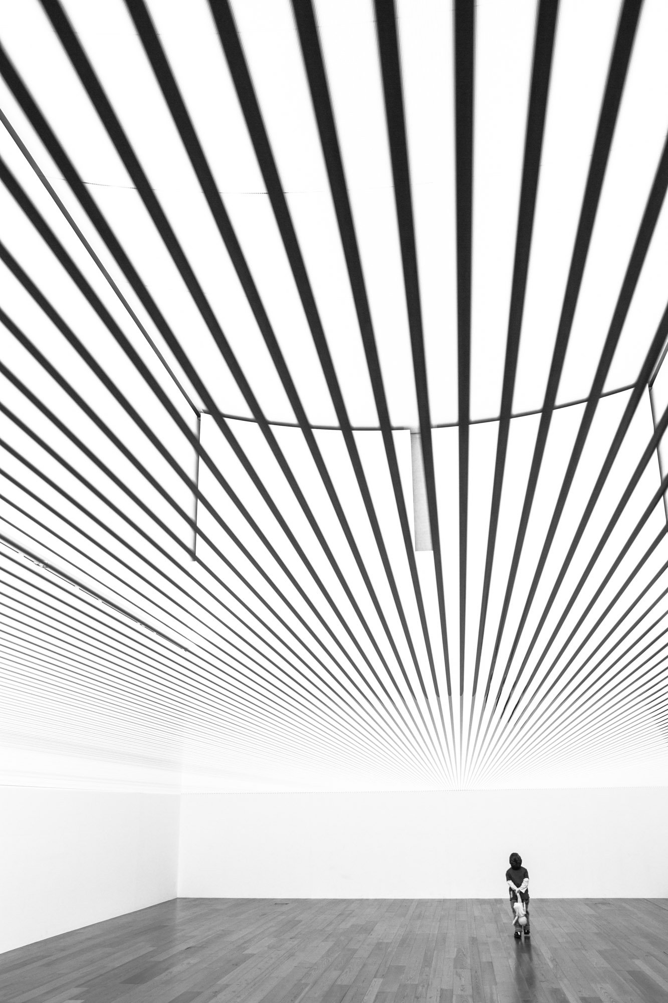A young child riding a scooter in a minimalist art gallery with black and white striped artwork on the wall.