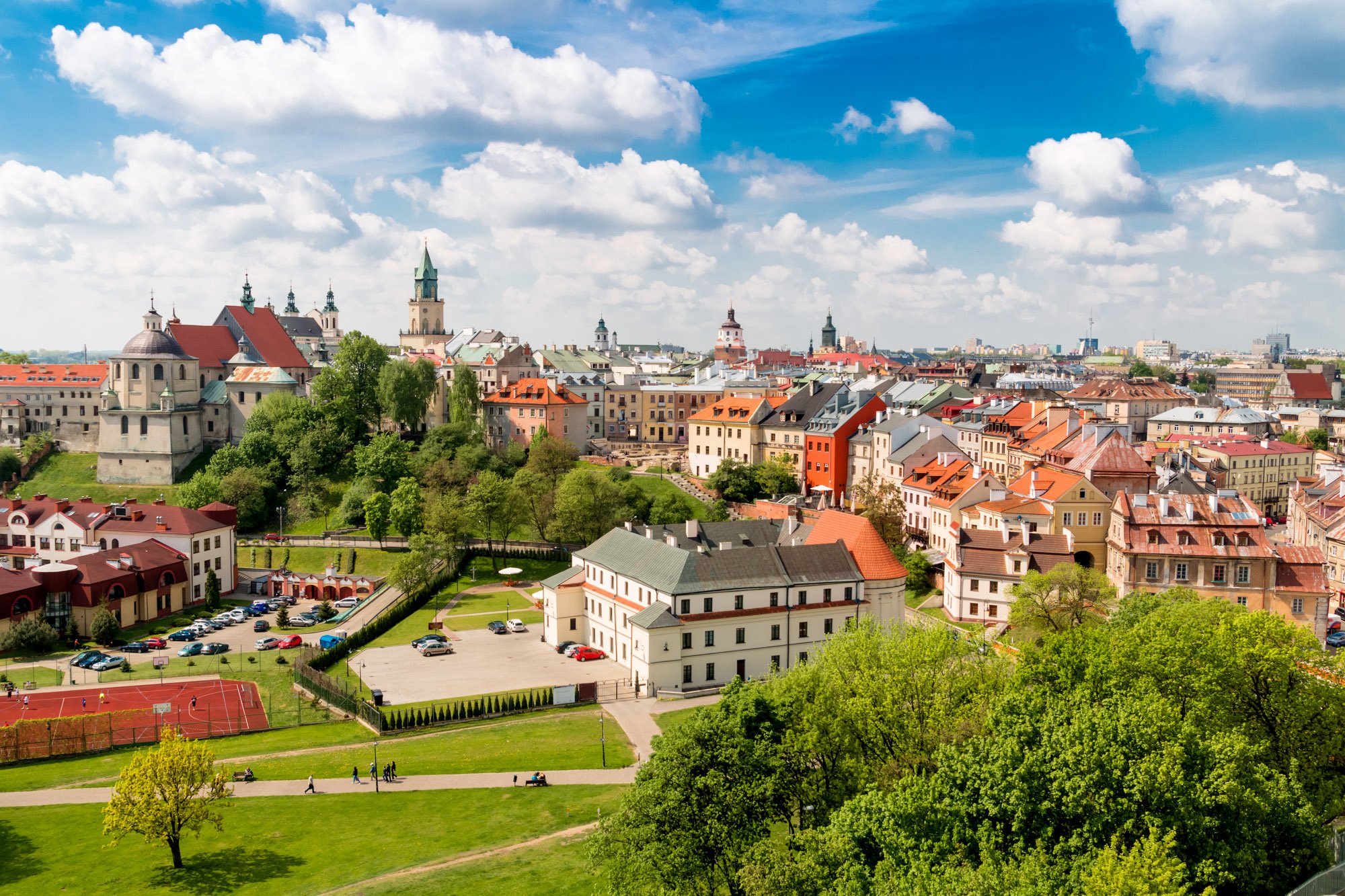 A panoramic view of a European city with colorful historic buildings, church steeples, and a lush green park with walking paths and benches under a partly cloudy sky.