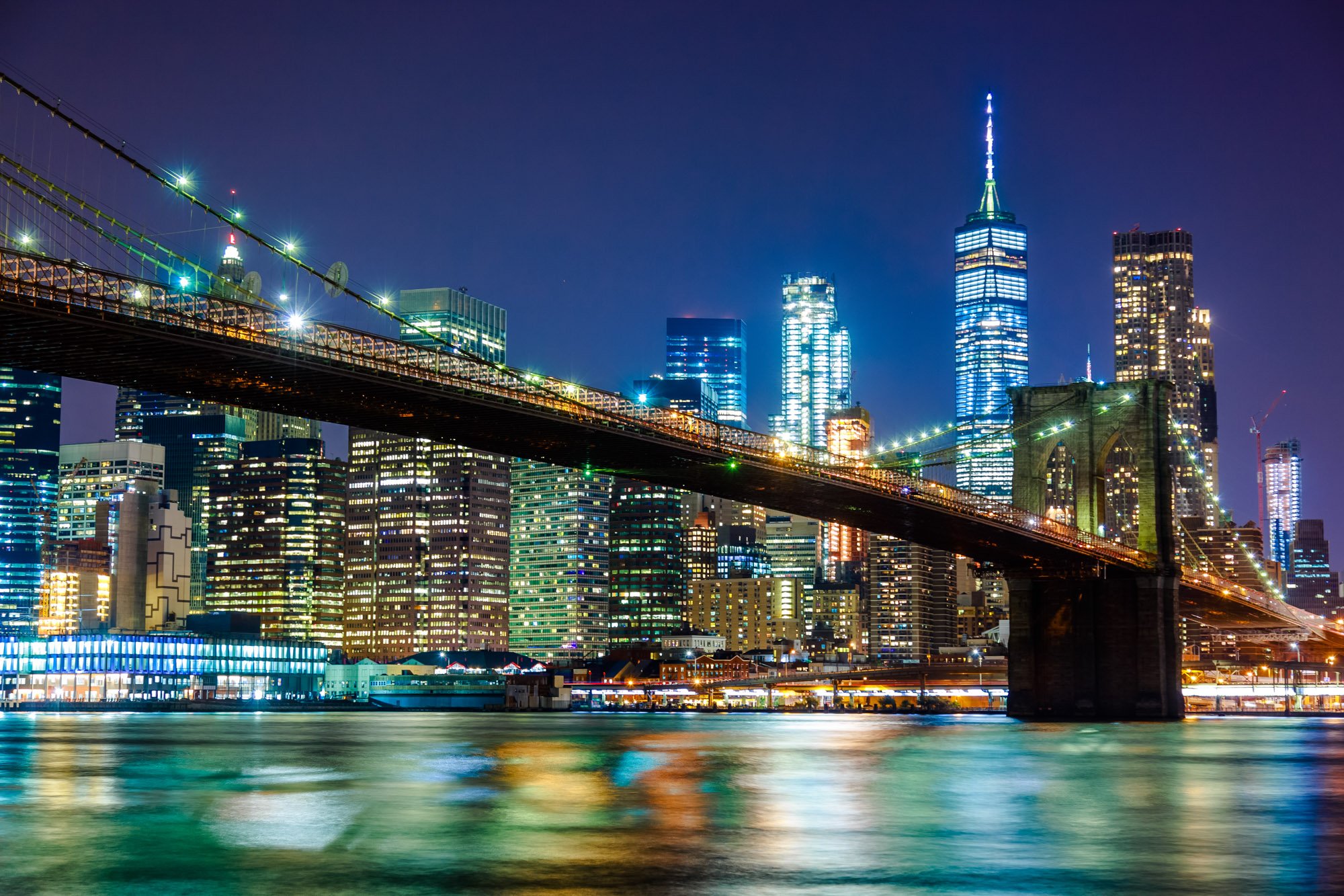 Night view of New York City skyline including the One World Trade Center lit up, buildings with illuminated windows, and the Brooklyn Bridge with lights reflected on the water.