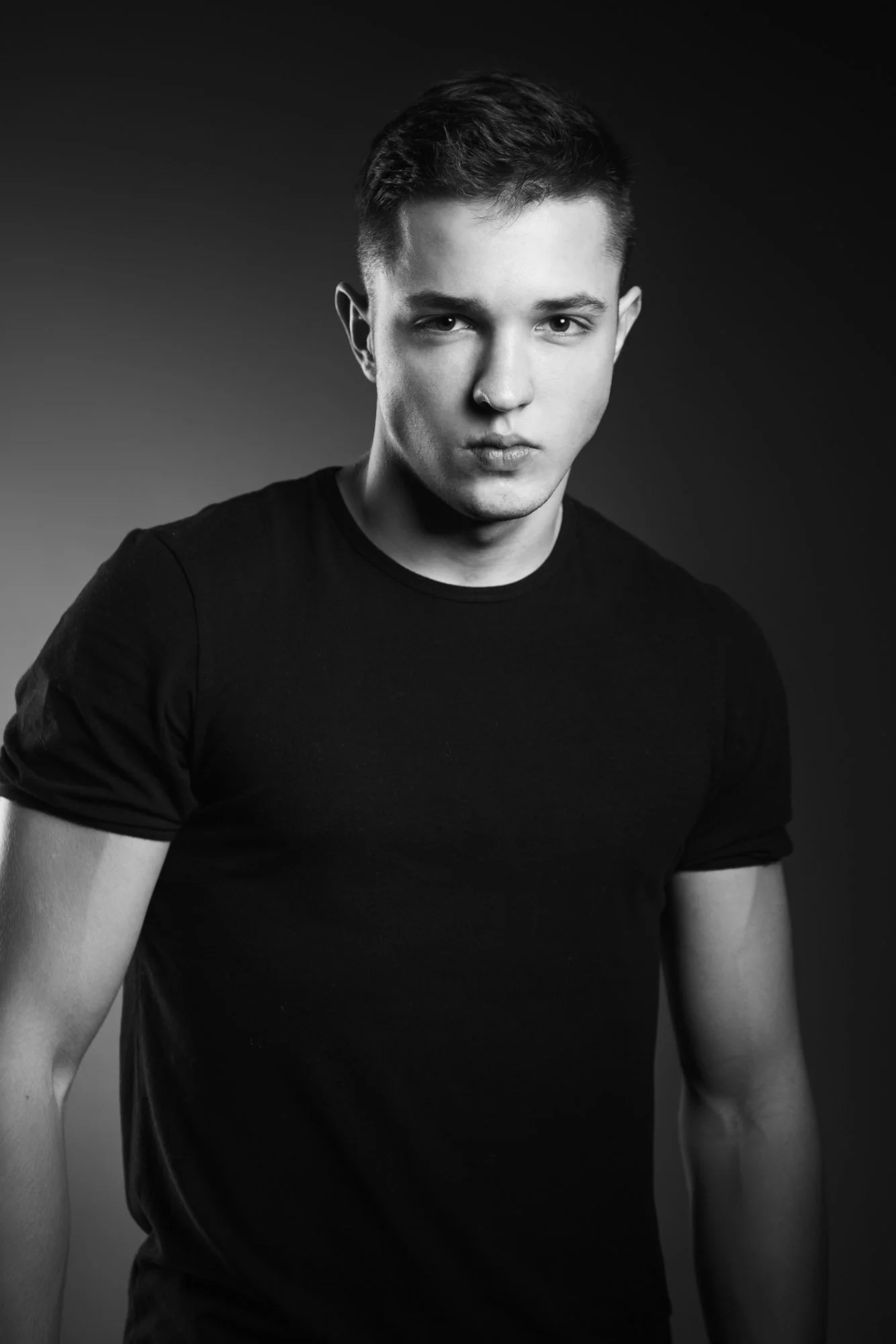 Black and white portrait of a young man with short dark hair, wearing a black t-shirt, looking directly at the camera with a serious expression against a gradient background.