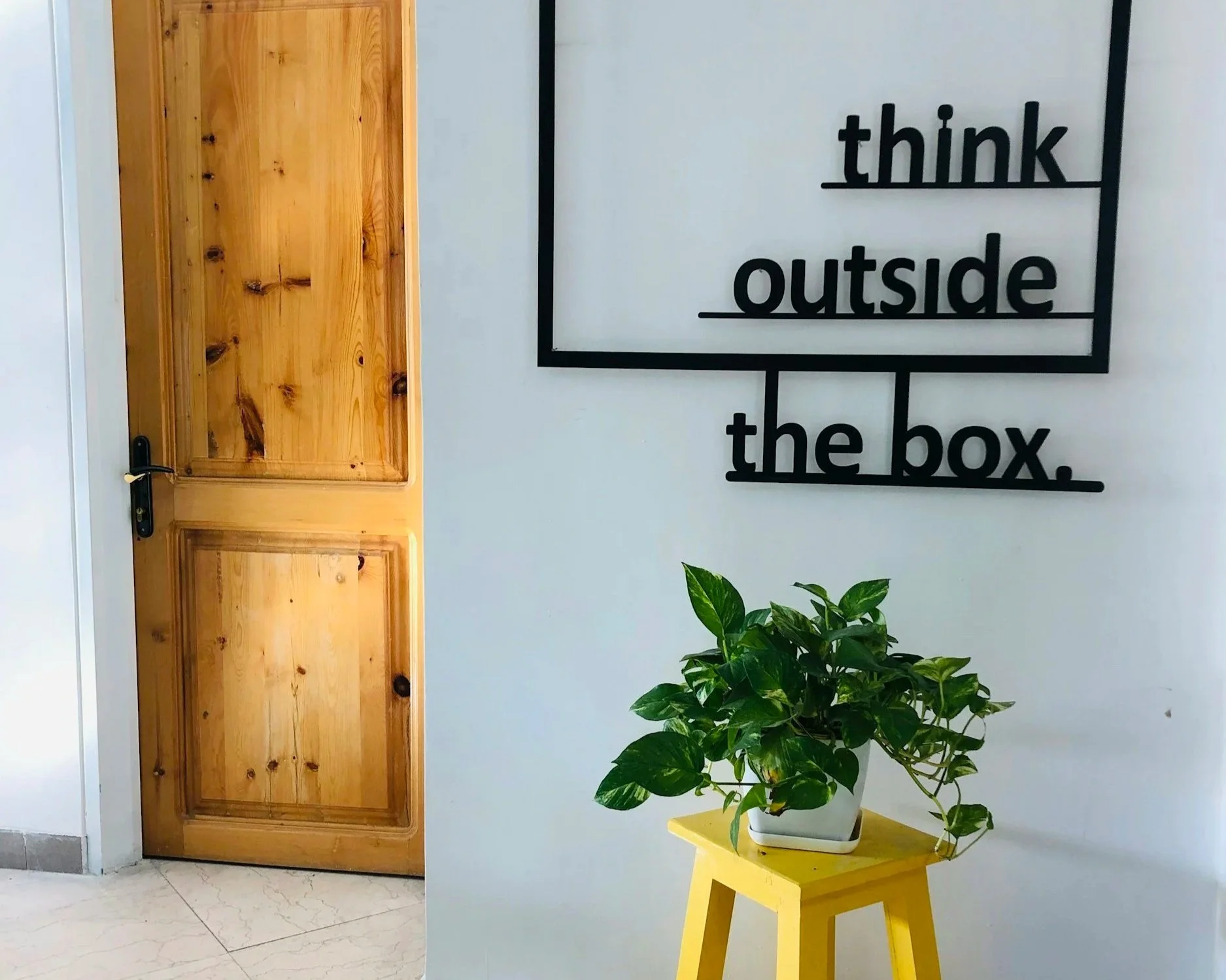 Corporate wall that says "think outside" within a square and "the box" outside of it, with a yellow stool holding a plant in the forefront and a door on the left side