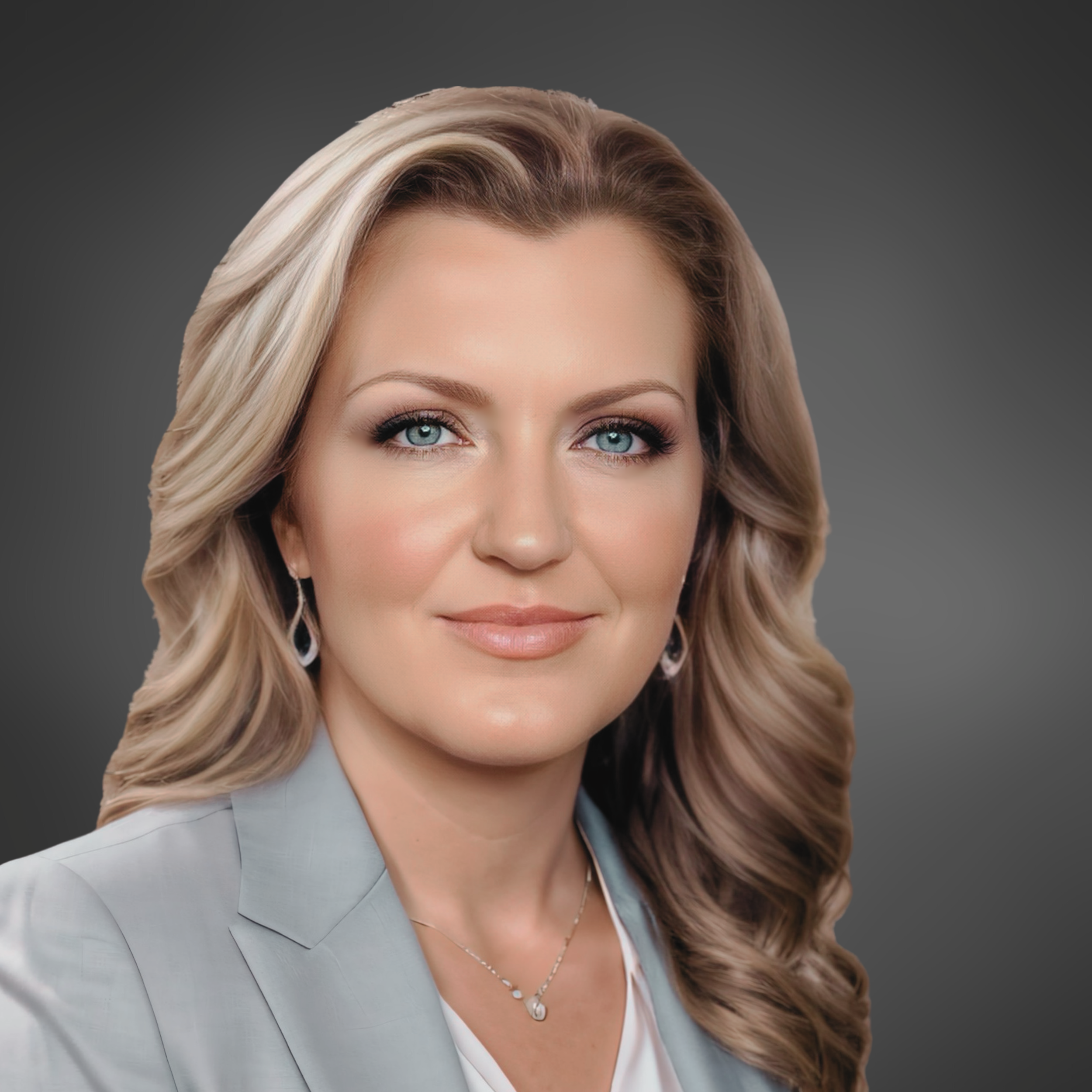 Professional headshot of a woman with long blonde hair and a light gray blazer, set against a smooth dark gray backdrop.