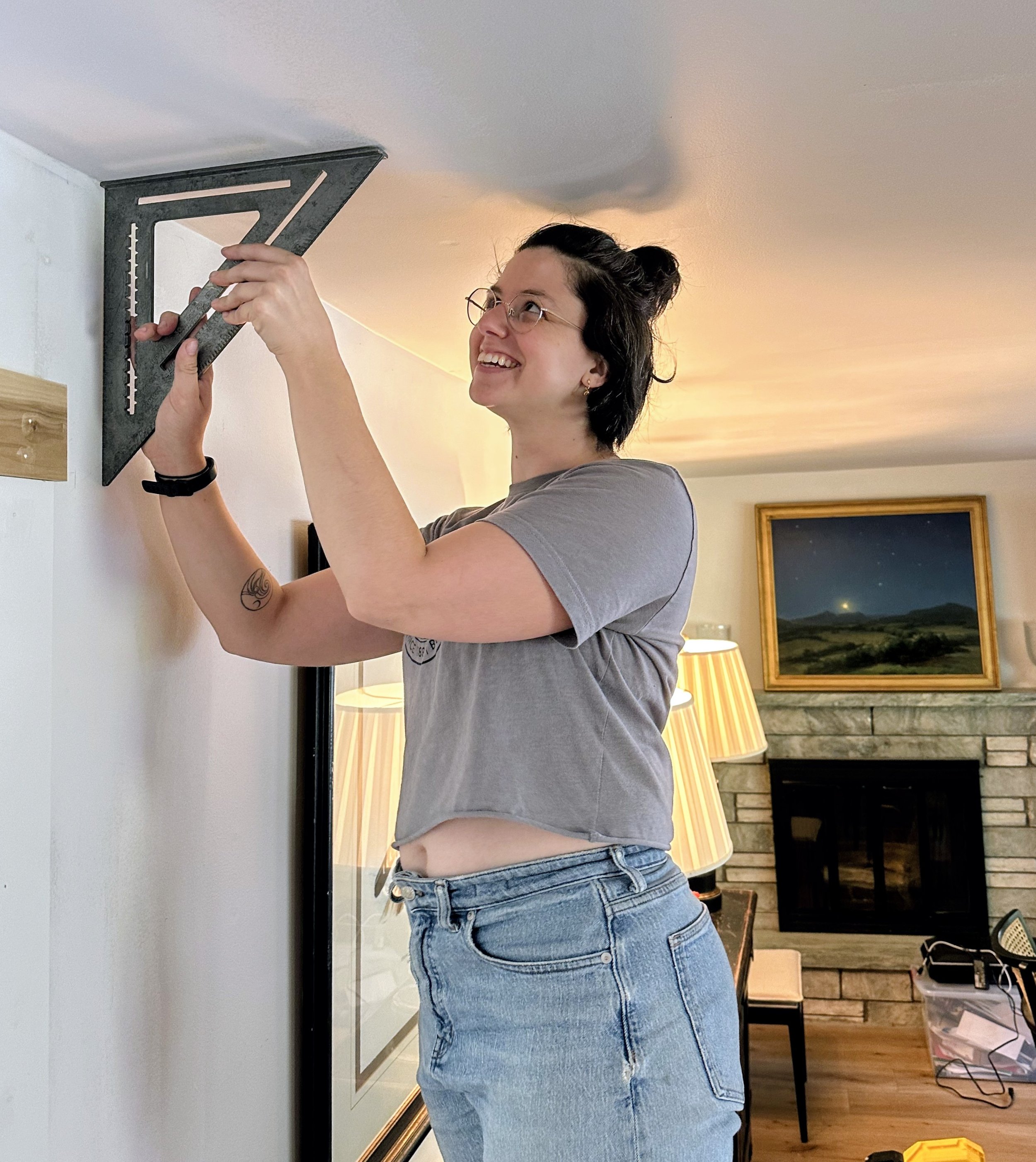 A woman with glasses and short dark hair installing a picture hanging bracket on a wall, smiling and looking up.