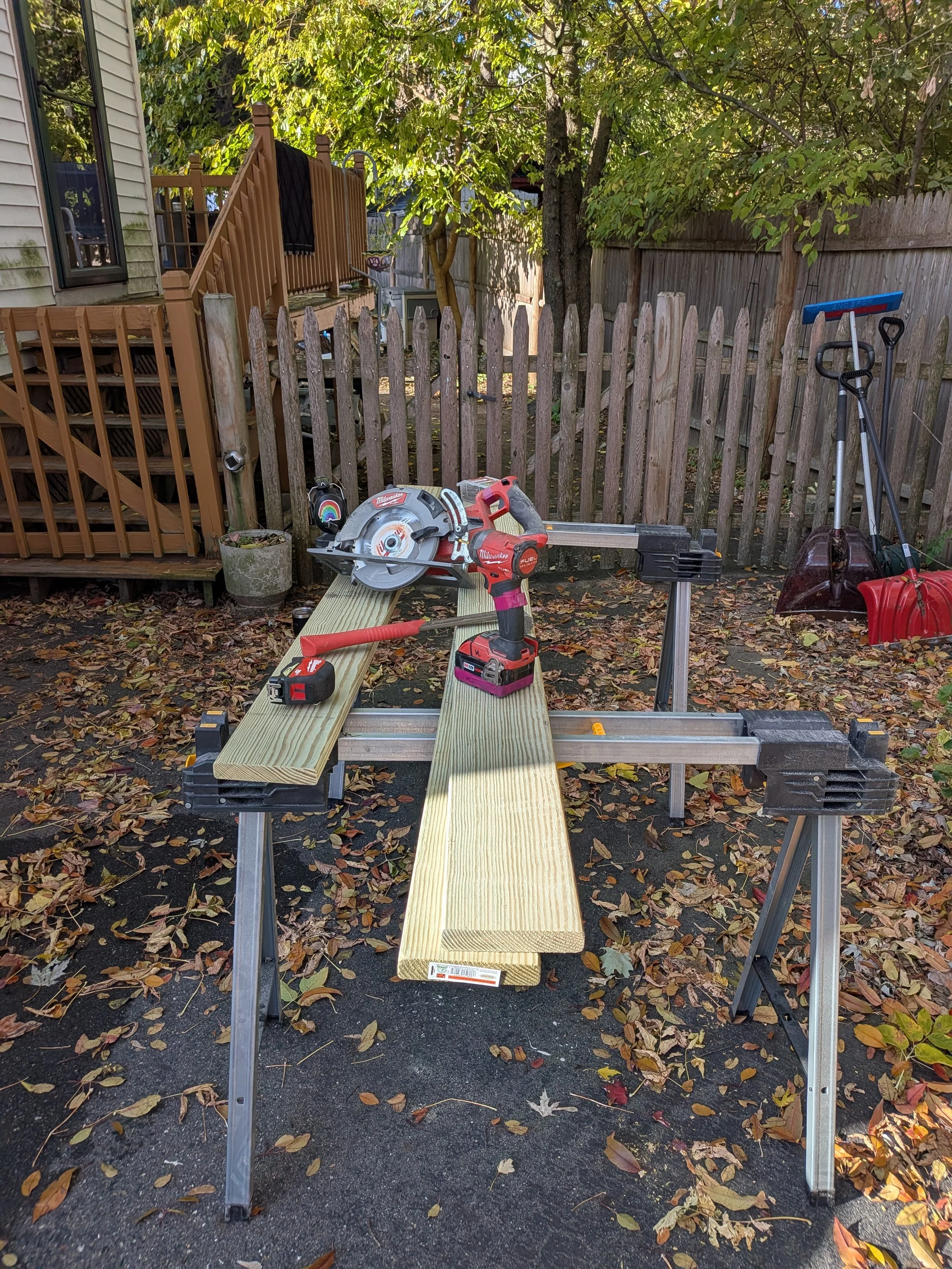 A woodworking workbench set up outdoors in a backyard, with various power tools and tools, surrounded by fallen autumn leaves, a wooden fence, trees, and a house with a staircase.