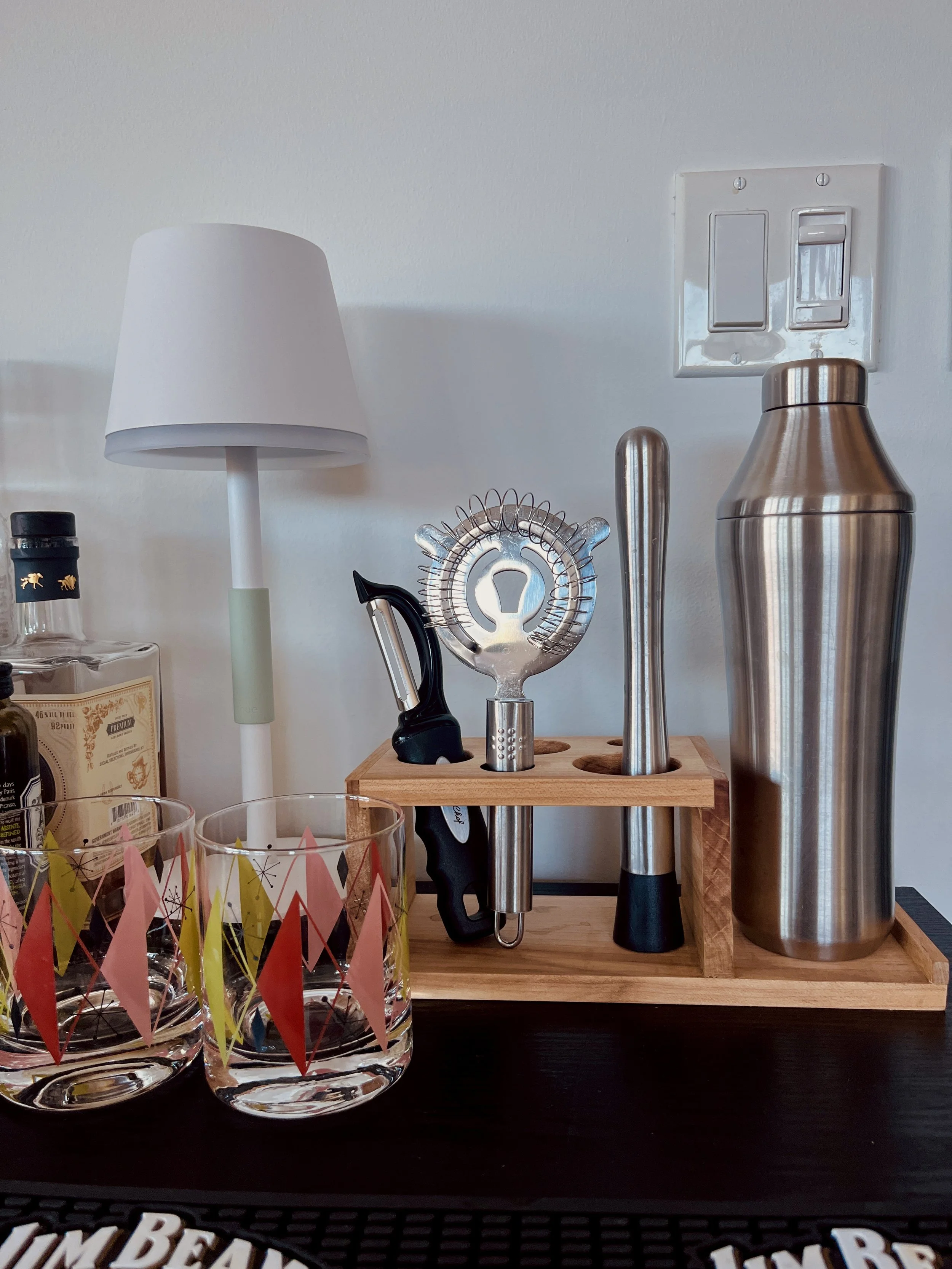 Bar cart with a small white lamp, cocktail shaker, bar tools in a wooden holder, glasses, and bottles against a white wall.