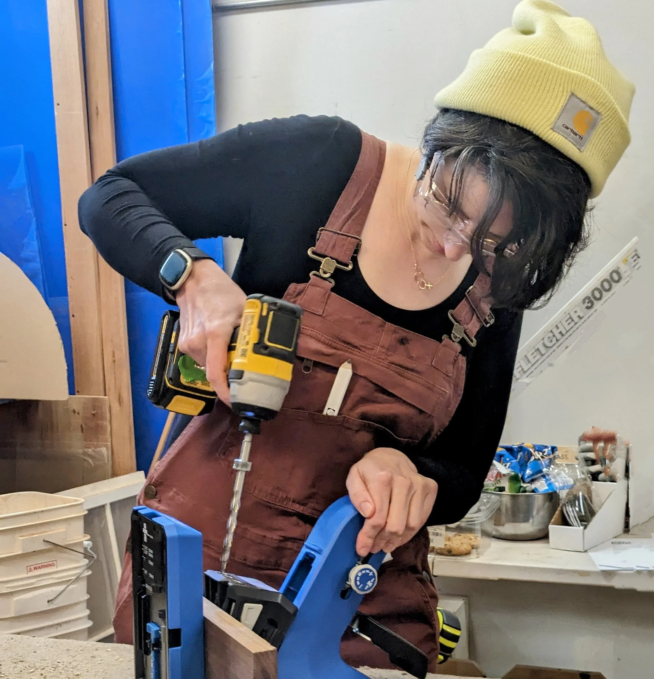Woman with black hair, glasses, and a yellow Carhartt beanie, wearing a black long-sleeve shirt and brown overalls, using a cordless drill on woodworking project in a workshop.