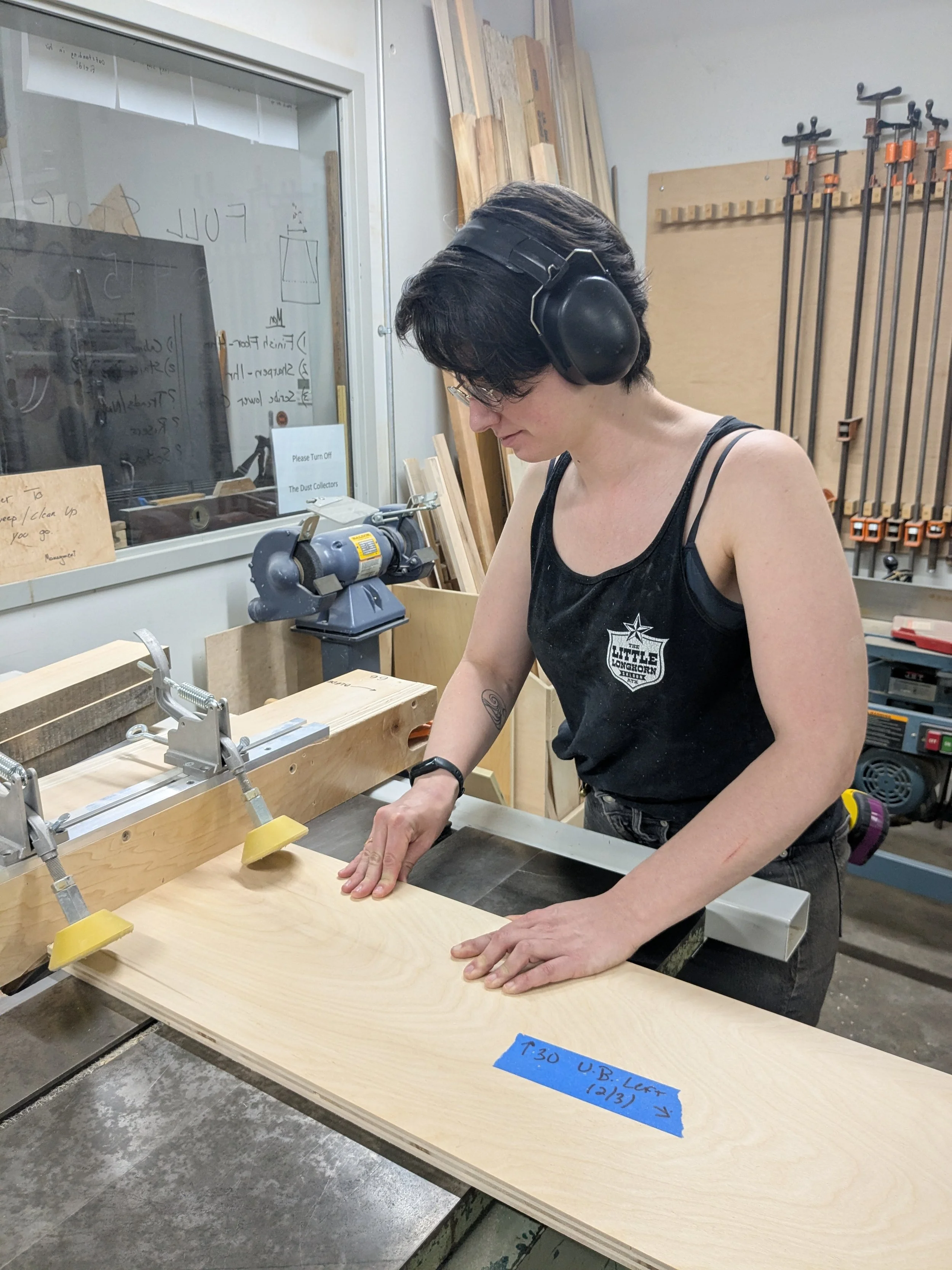 A woman wearing glasses and protective headphones working on a woodworking project using a clamp and a saw in a woodworking shop.