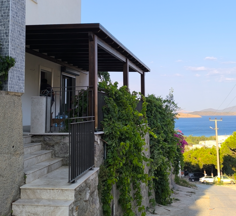 Çatı altındaki veranda, merdivenlerle çıkış ve yeşil bitkilerle çevrili, deniz ve ağaçlarla dolu manzara görünümü.