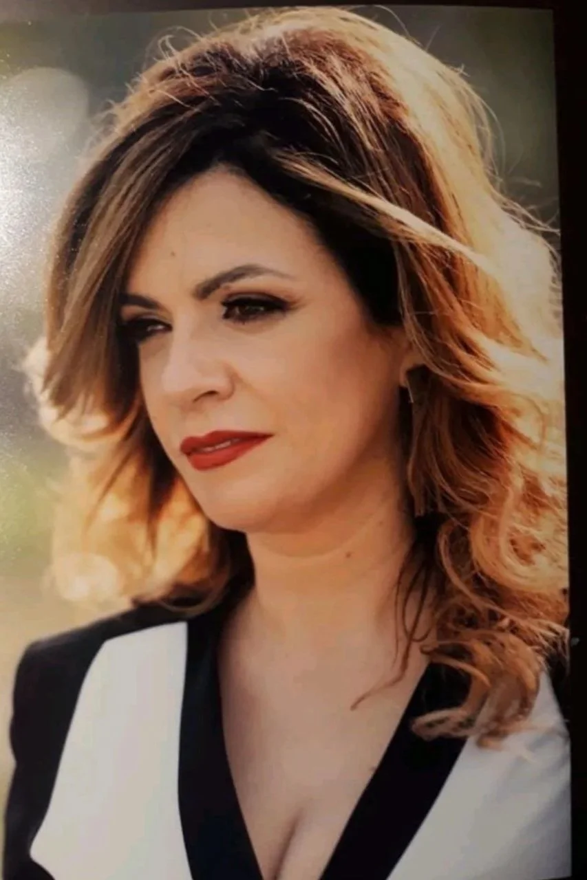 Close-up of a woman with shoulder-length wavy hair and red lipstick, wearing a black and white outfit, outdoors with blurred background.