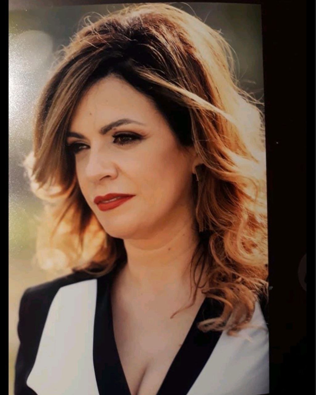 A woman with shoulder-length wavy brown hair with highlights, wearing red lipstick and a black and white top, outdoors in natural light.