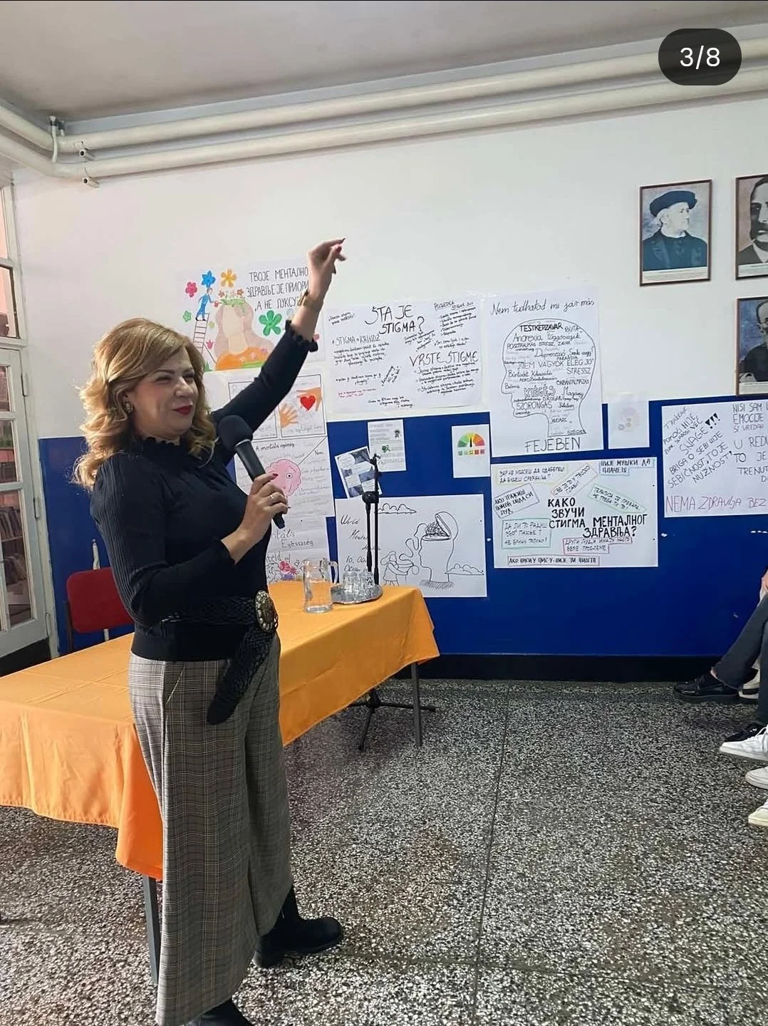 A woman standing at a presentation, holding a microphone and gesturing with her hand, in front of a display with various posters and notes about mental health and stigma, in a classroom or conference room setting.