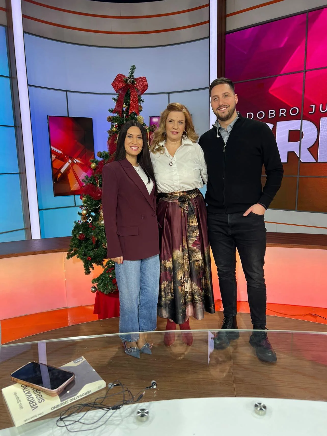 Three people, two women and one man, standing together in a TV studio with a decorated Christmas tree behind them. The woman on the left is wearing a maroon blazer and jeans, the woman in the middle is wearing a white blouse and a patterned skirt, and the man on the right is wearing a black jacket and dark pants. There is a smartphone, a book, and some wired earbuds on a glass table in the foreground.
