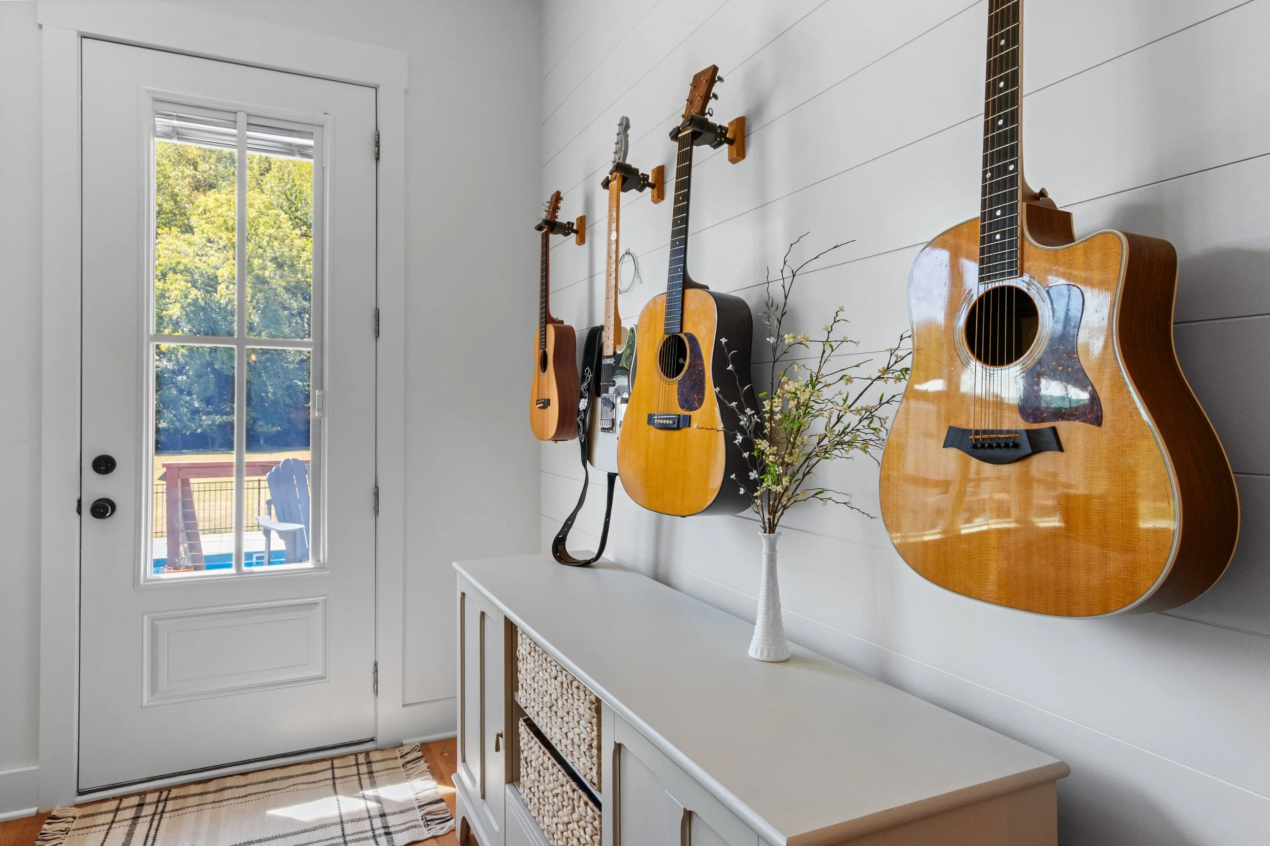 Four guitars hanging on a white wall, with a white cabinet below and a vase with branches and flowers on top. A glass door with a view of a deck and trees outside.