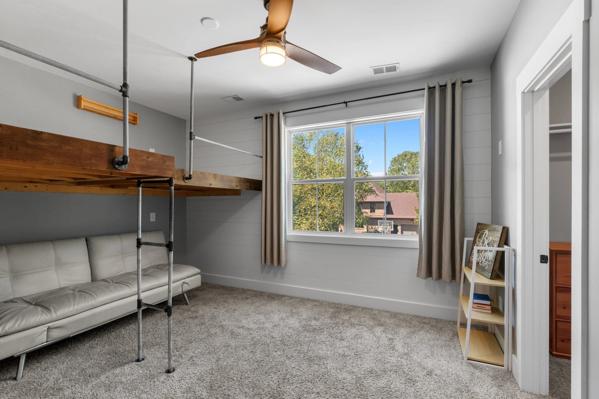A room with a large window showing trees and a building outside, a white leather sofa, beige curtains, a ceiling fan, a small white bookshelf with decorative items, and a loft with a wooden guardrail.