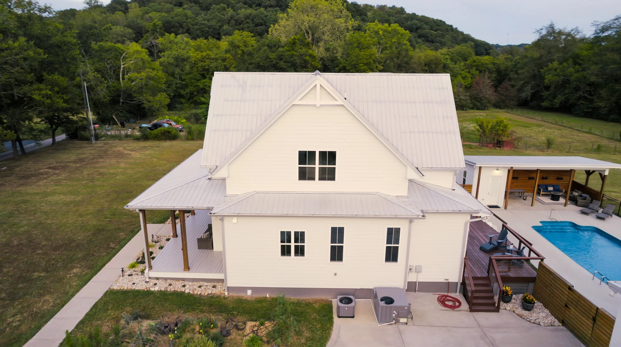 A white two-story house with a metal roof, surrounded by a lawn and trees, featuring a backyard with a pool, a covered seating area, and a wooden deck.