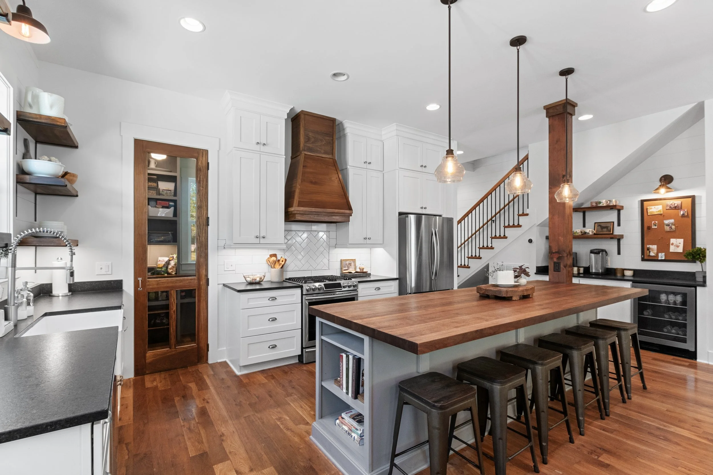 Spacious kitchen with white cabinets, a wooden island with stools, a stainless steel refrigerator, and a staircase in the background.
