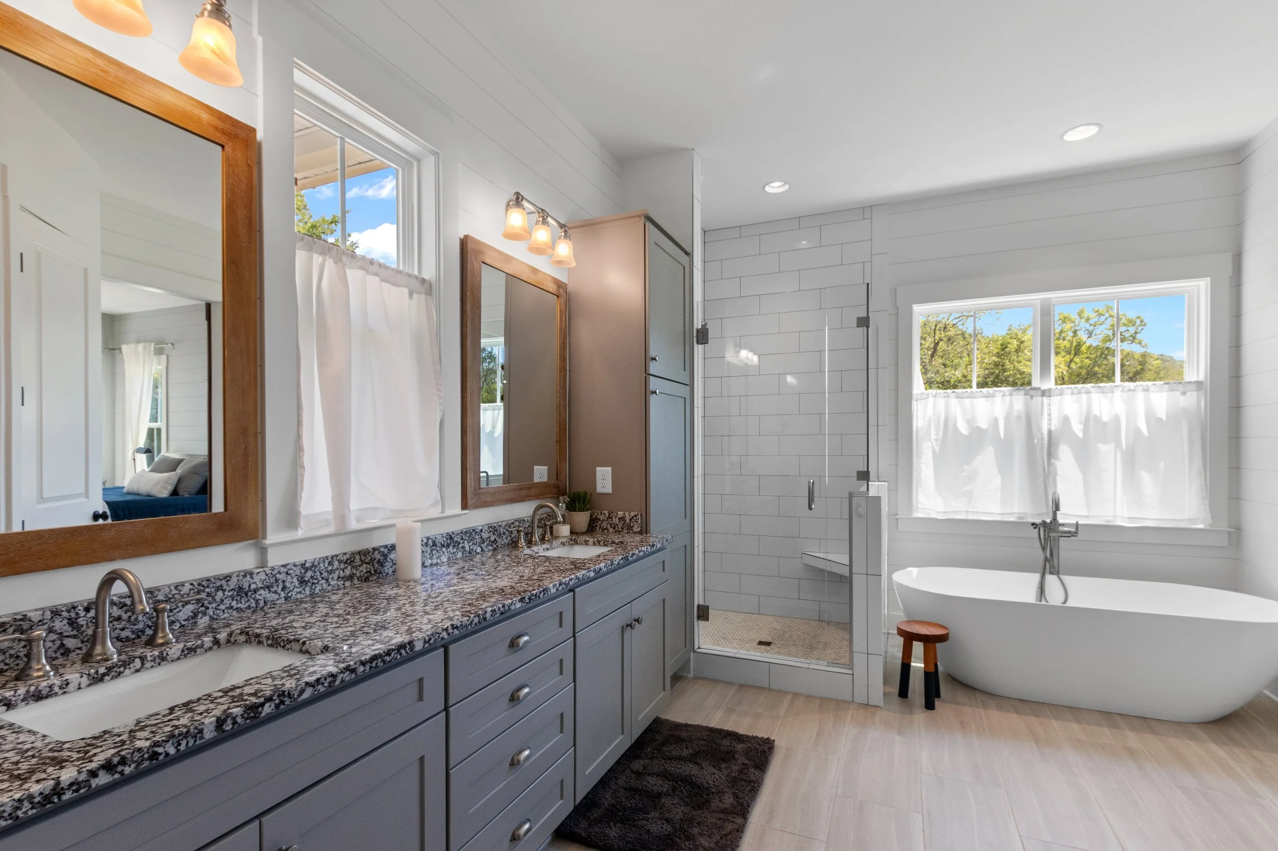 Bright bathroom with dual sinks, granite countertops, framed mirrors, a built-in cabinet, and a walk-in shower with glass door. There's a freestanding tub under a large window with white curtains. Wooden accents and light-colored tiles add warmth to