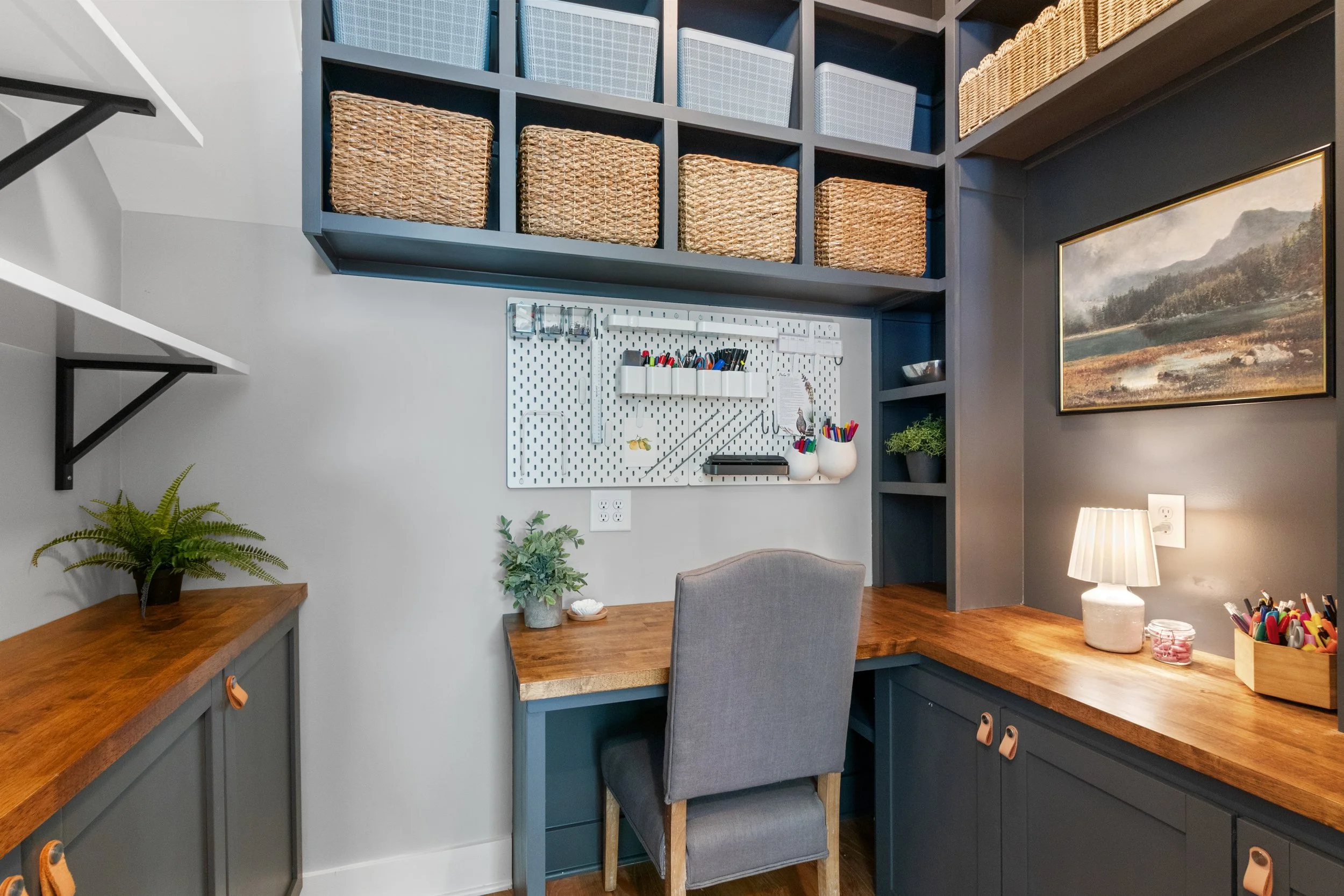 Home office corner with a gray upholstered chair, wooden desk, blue cabinetry, built-in shelves, storage baskets, a framed landscape painting, a small table lamp, potted plants, and a pegboard with office supplies.