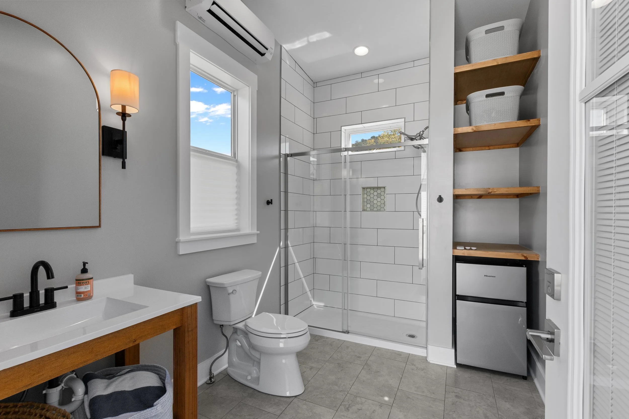 Modern bathroom with white tiled shower, grey walls, wooden shelves with baskets, small refrigerator, white sink with black fixtures, toilet, window, air conditioning unit, and a wall-mounted light.