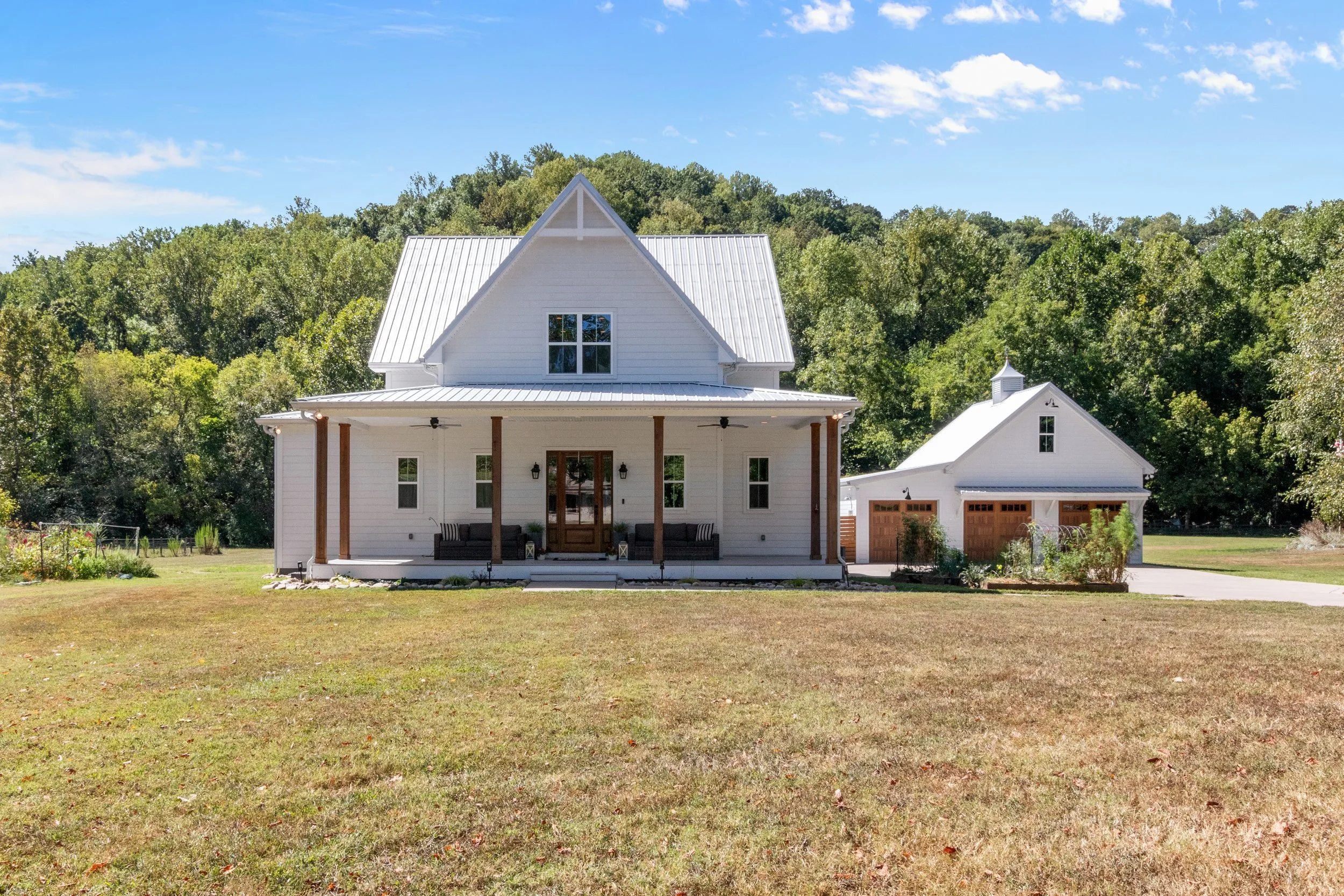 White farmhouse-style house with a front porch, front door, multiple windows, and a metal roof, set against a wooded backdrop with a blue sky and a detached garage with wooden doors to the right.