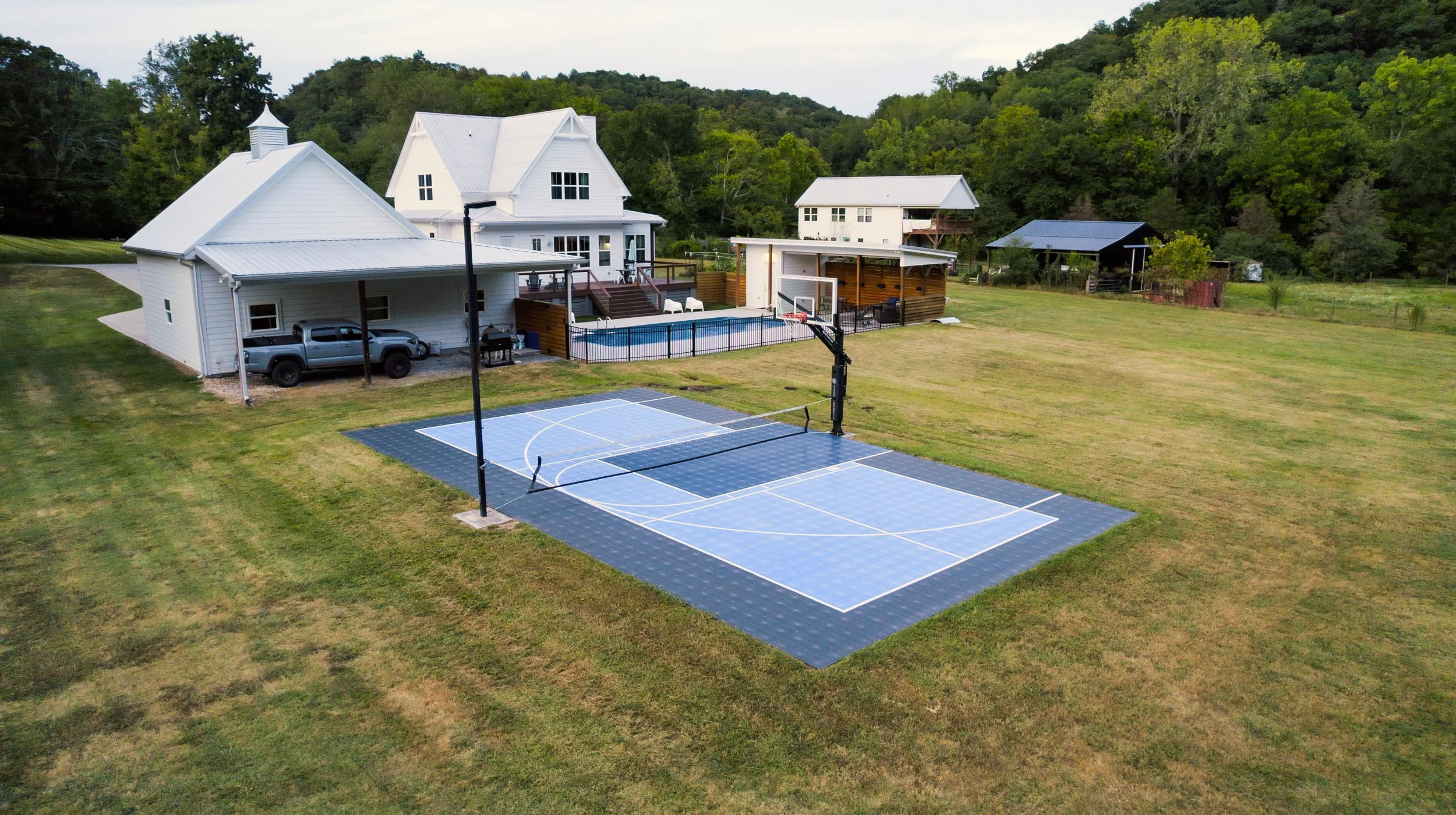 A backyard with a basketball court, a swimming pool, and neighboring houses with a wooded hillside in the background.