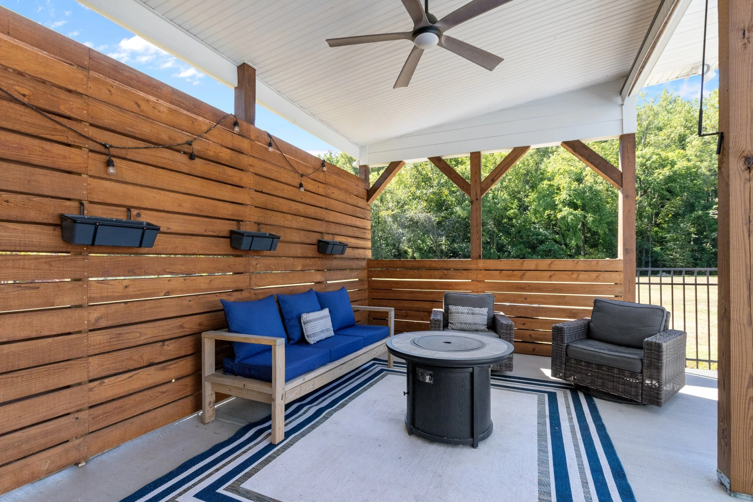 Covered outdoor patio with wooden privacy wall, ceiling fan, string lights, wicker chairs, a wooden bench with blue cushions, a round firepit table, and a striped outdoor rug, overlooking green trees and an open field.