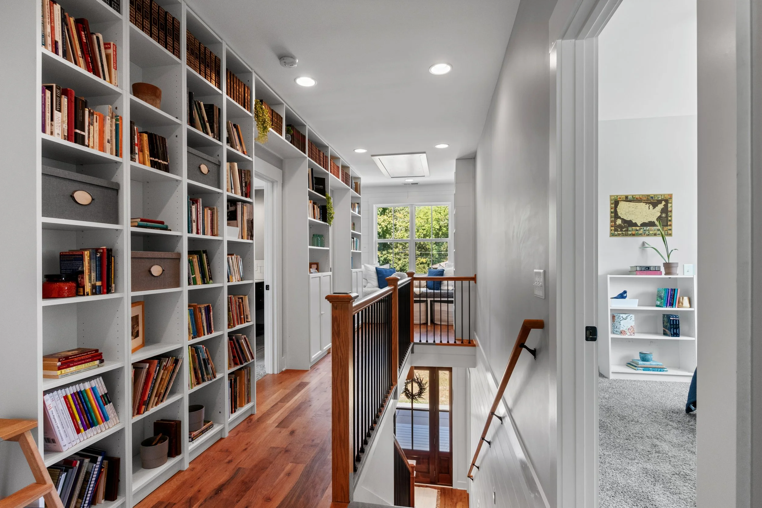 Bright upper-floor hallway with white walls, hardwood floor, built-in white bookshelves filled with books and decor, a staircase with a wooden handrail, and two rooms visible through open doors; large window at the end of the hall with greenery outsi