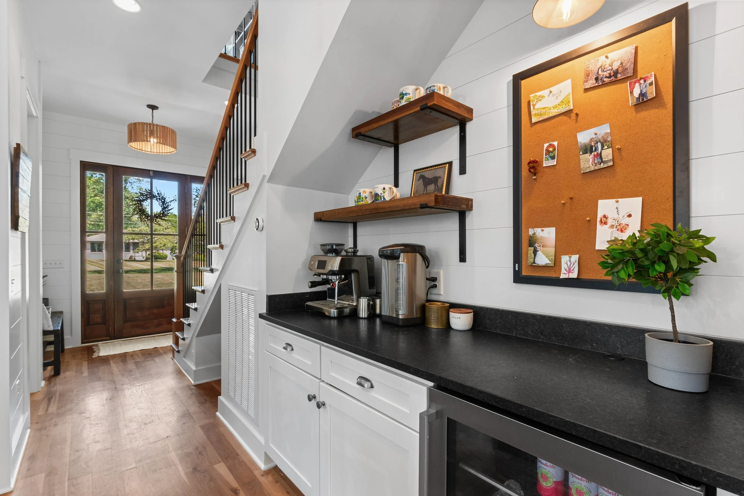 Interior of a modern home kitchen near the back entrance with a countertop, coffee maker, and coffee dispenser. Shelves above hold cups and decor, with a large corkboard displaying photos and artwork, and a potted plant nearby.