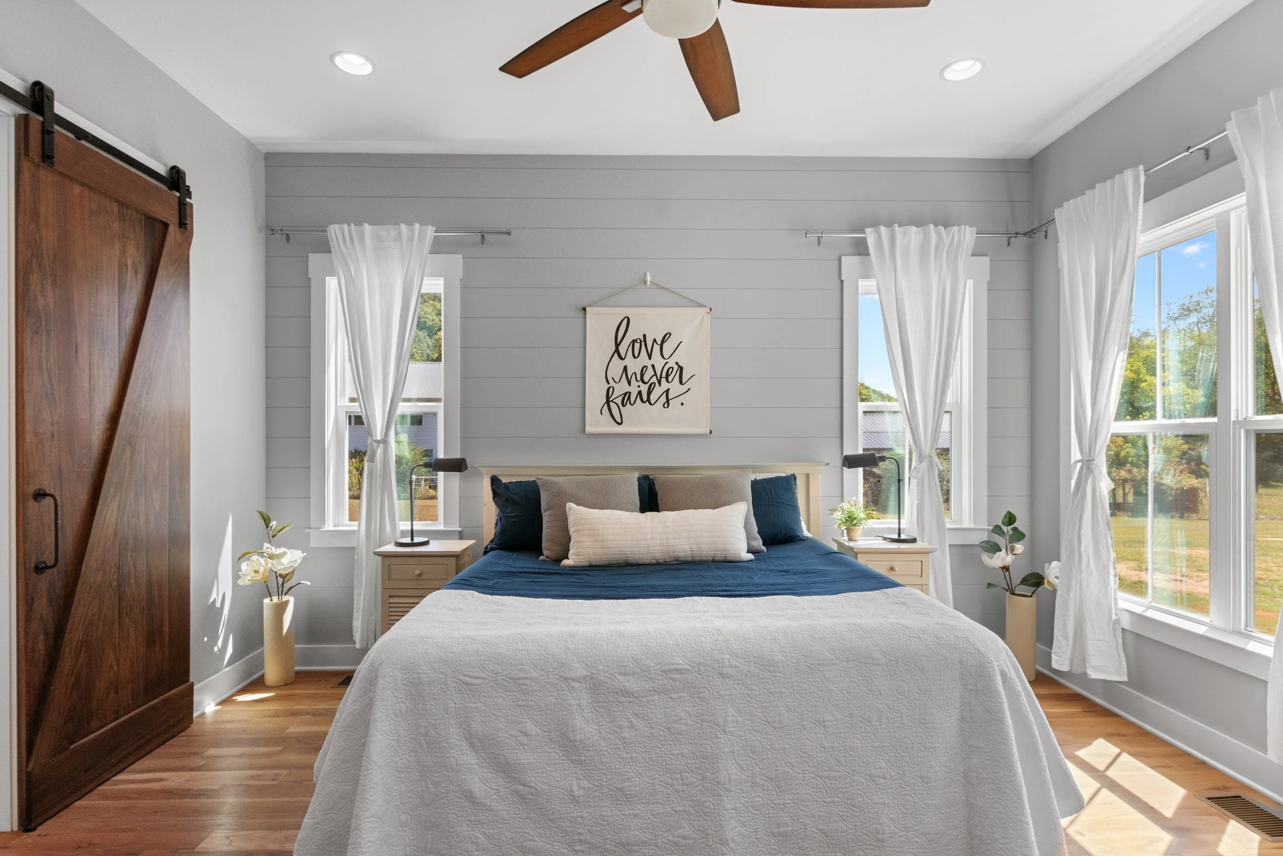 Bright bedroom with white walls, hardwood flooring, and large windows with white curtains. A bed with navy blue and beige pillows and a white quilt is centered in the room. Two nightstands with black lamps and small plants flank the bed. A wooden sli