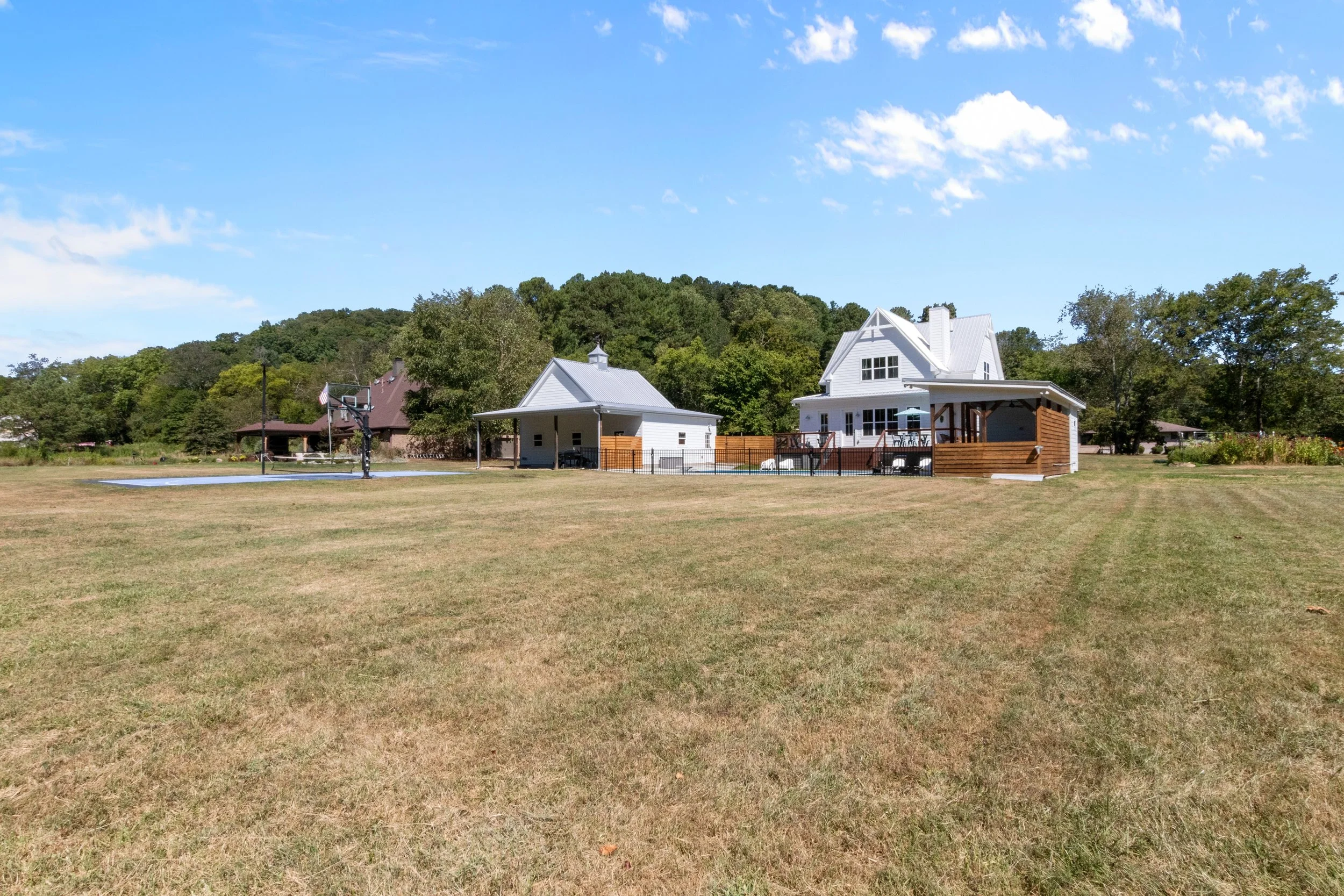 Large backyard with a house, a shed, and a basketball court under a blue sky with clouds.