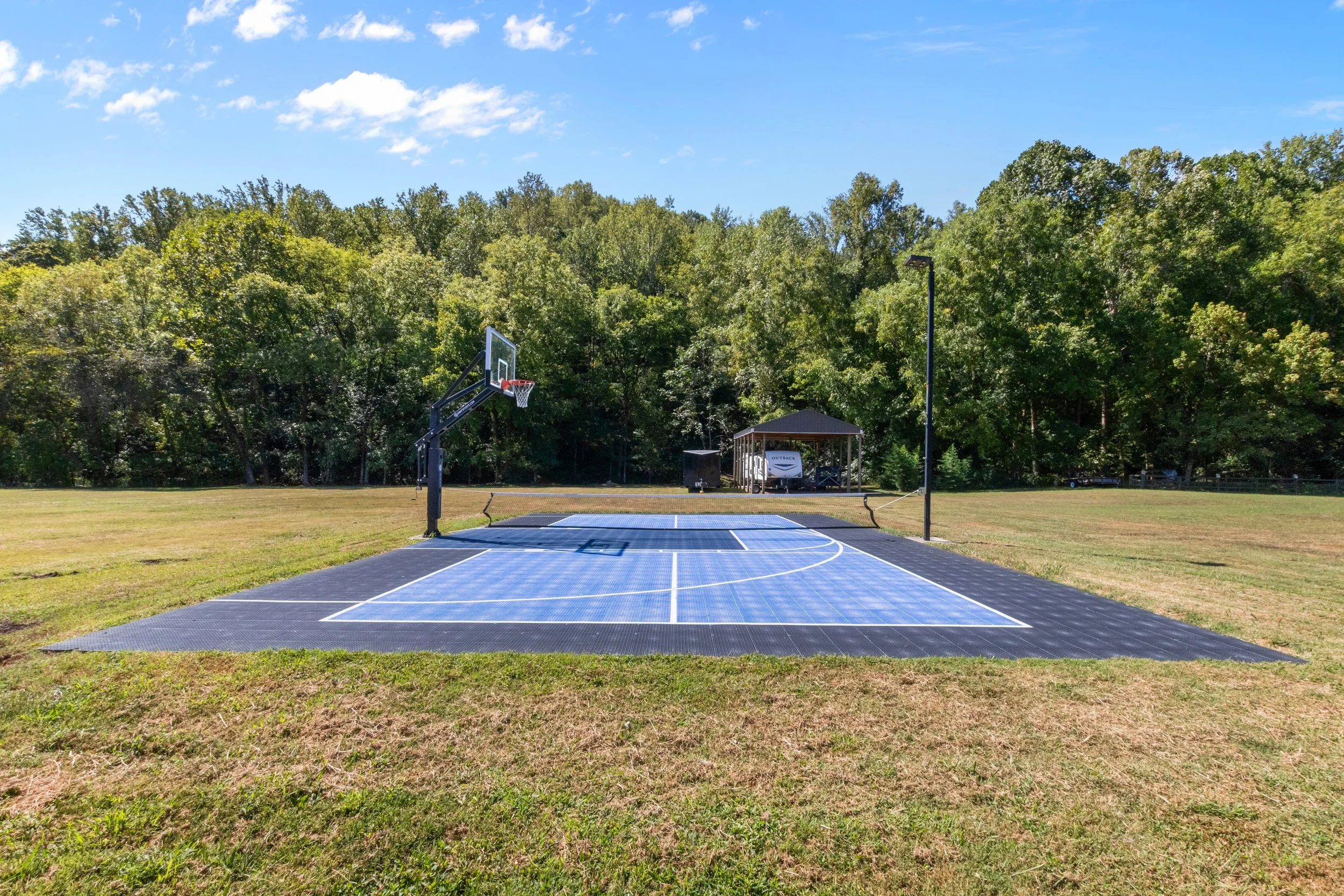 An outdoor basketball court with a solar panel surface, a hoop with a backboard, and trees in the background under a blue sky.