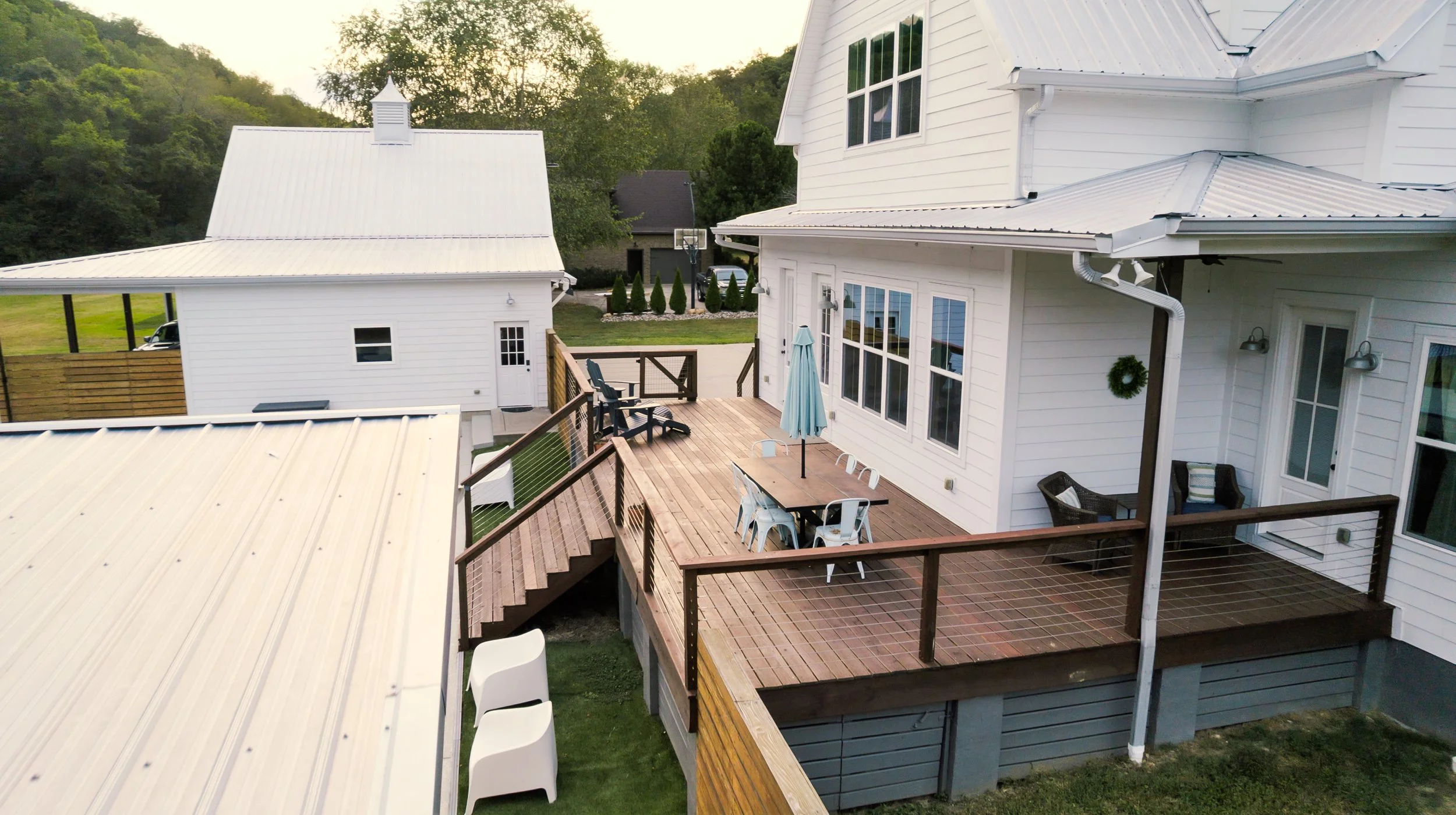 A large white house with a wooden deck outdoor patio, patio table with an umbrella, chairs, and two surrounding chairs with cushions. There is a smaller white building and fenced area in the background, surrounded by green trees and hills.