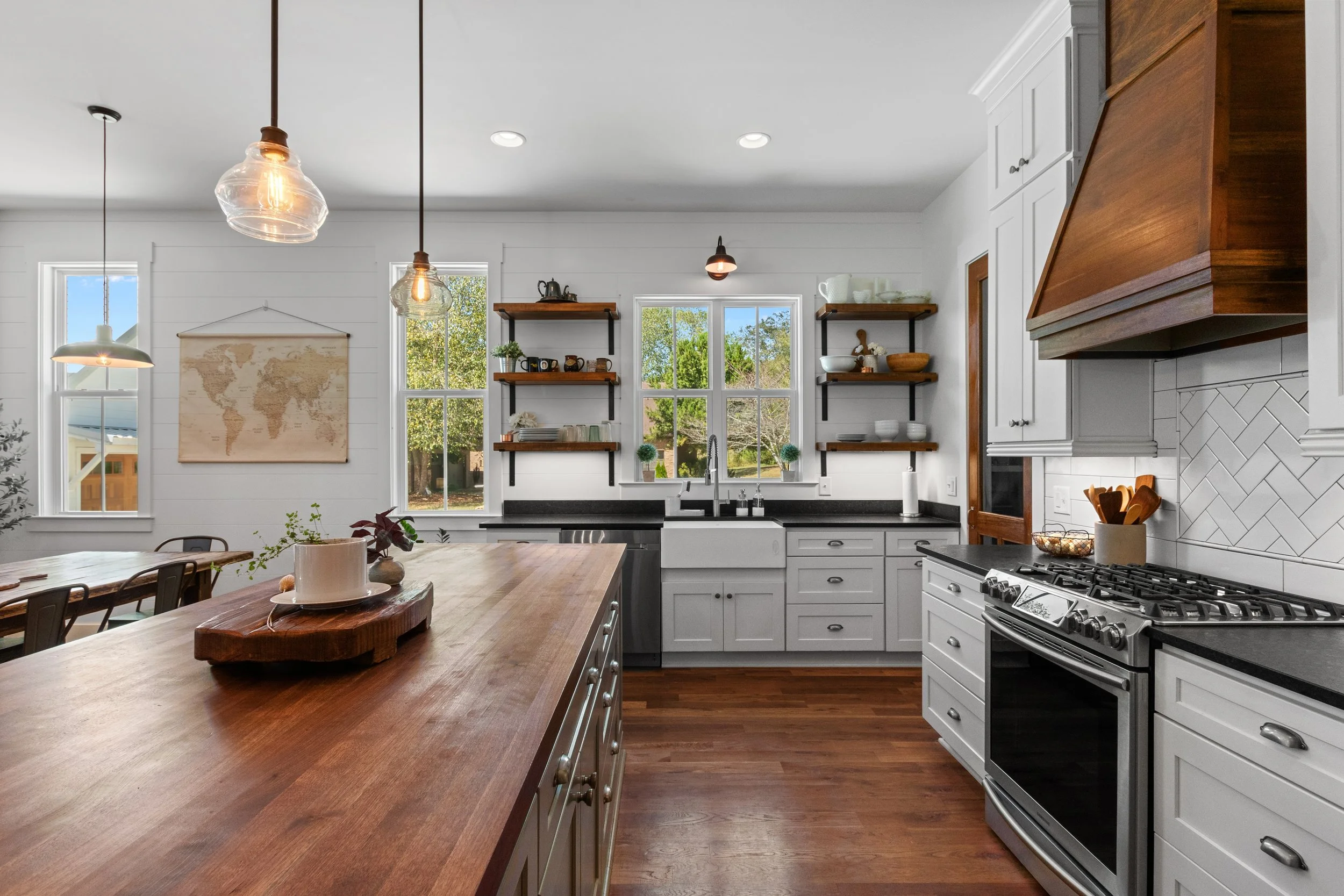 Modern kitchen with white cabinets, black countertops, wooden open shelves, stainless steel stove, farmhouse sink, and pendant lights, overlooking a backyard with trees