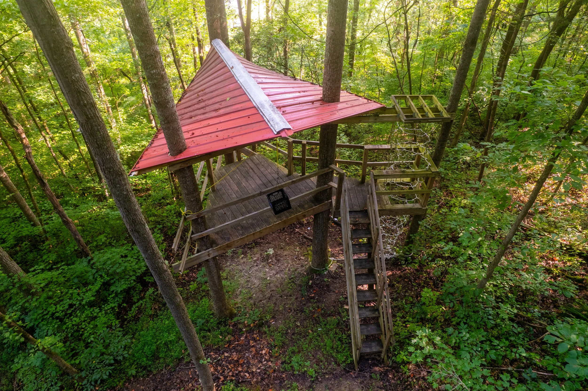 A treehouse with a red metal roof built among tall trees in a forest, featuring a wooden deck, stairs, and platforms with safety railings.