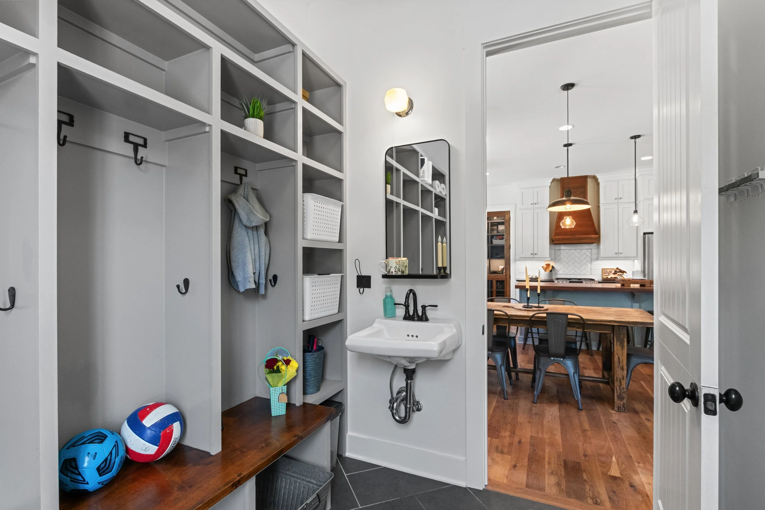Mudroom with built-in cubbies and hooks, small sink with mirror and decorations, chairs with sports balls placed on a wooden bench, leading to a dining kitchen with wooden table and chairs.